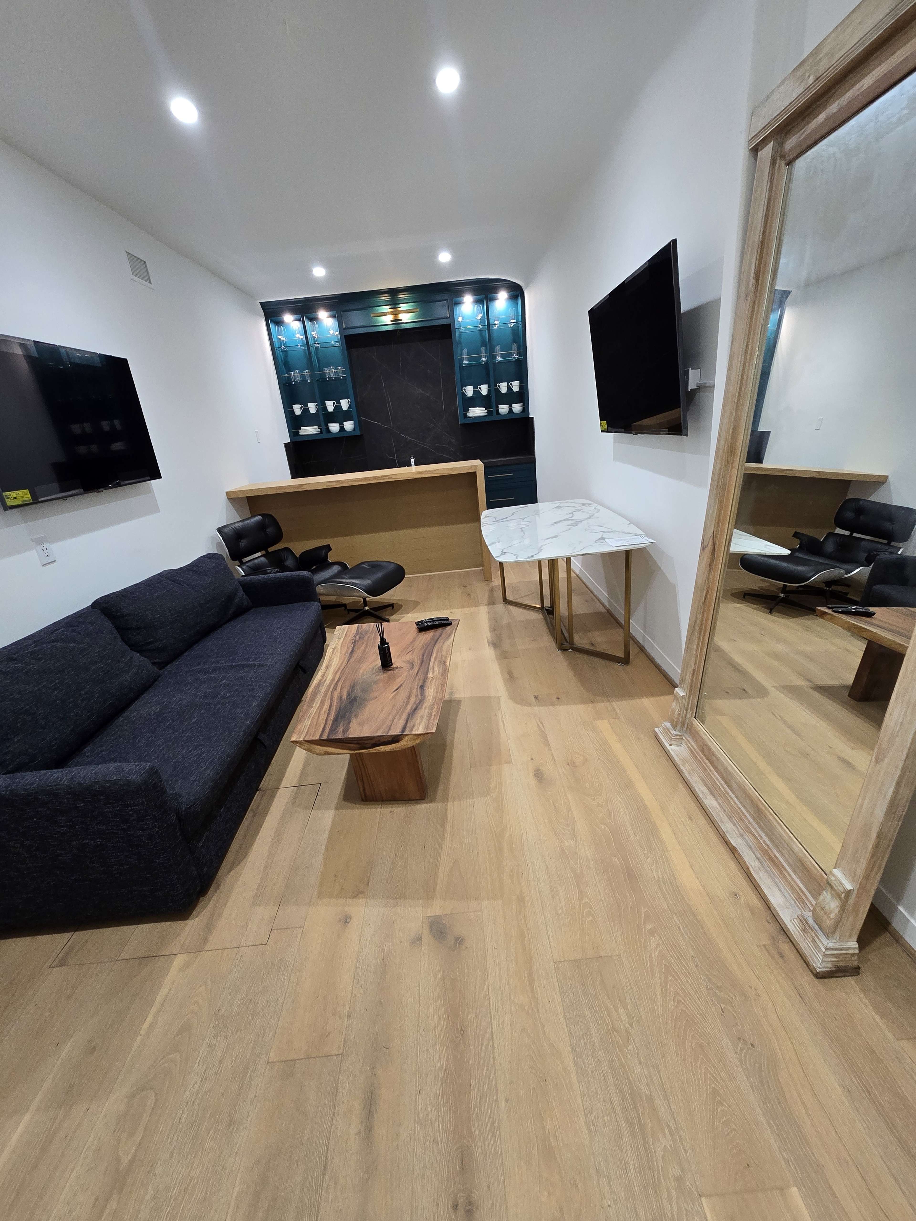 The room features a modern lounge area with a dark sofa, a wooden coffee table, two flat-screen TVs mounted on the walls, and a large mirror reflecting the space.