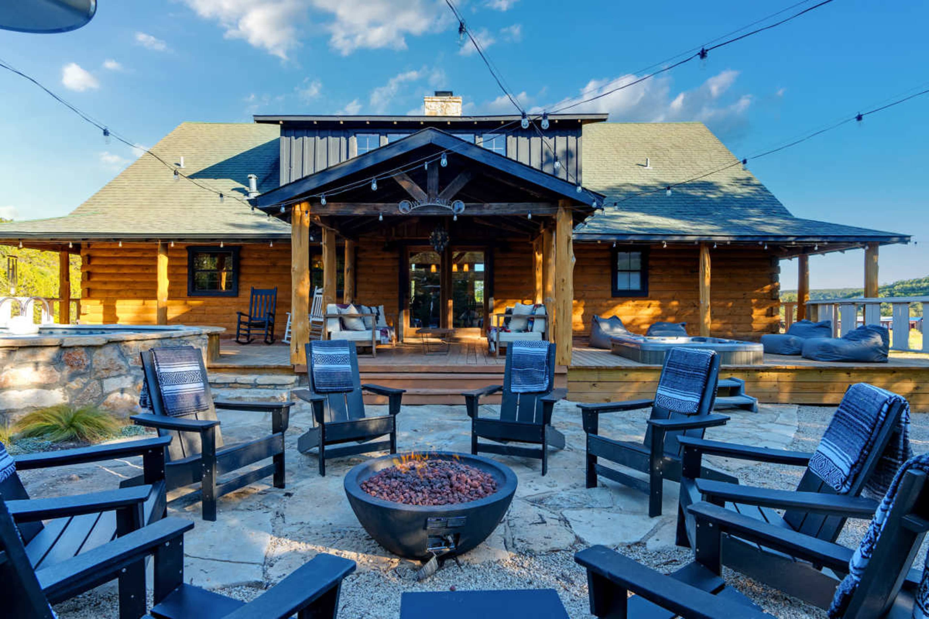 The image shows a log cabin with a porch, surrounded by several black chairs arranged around a fire pit on a stone patio.
