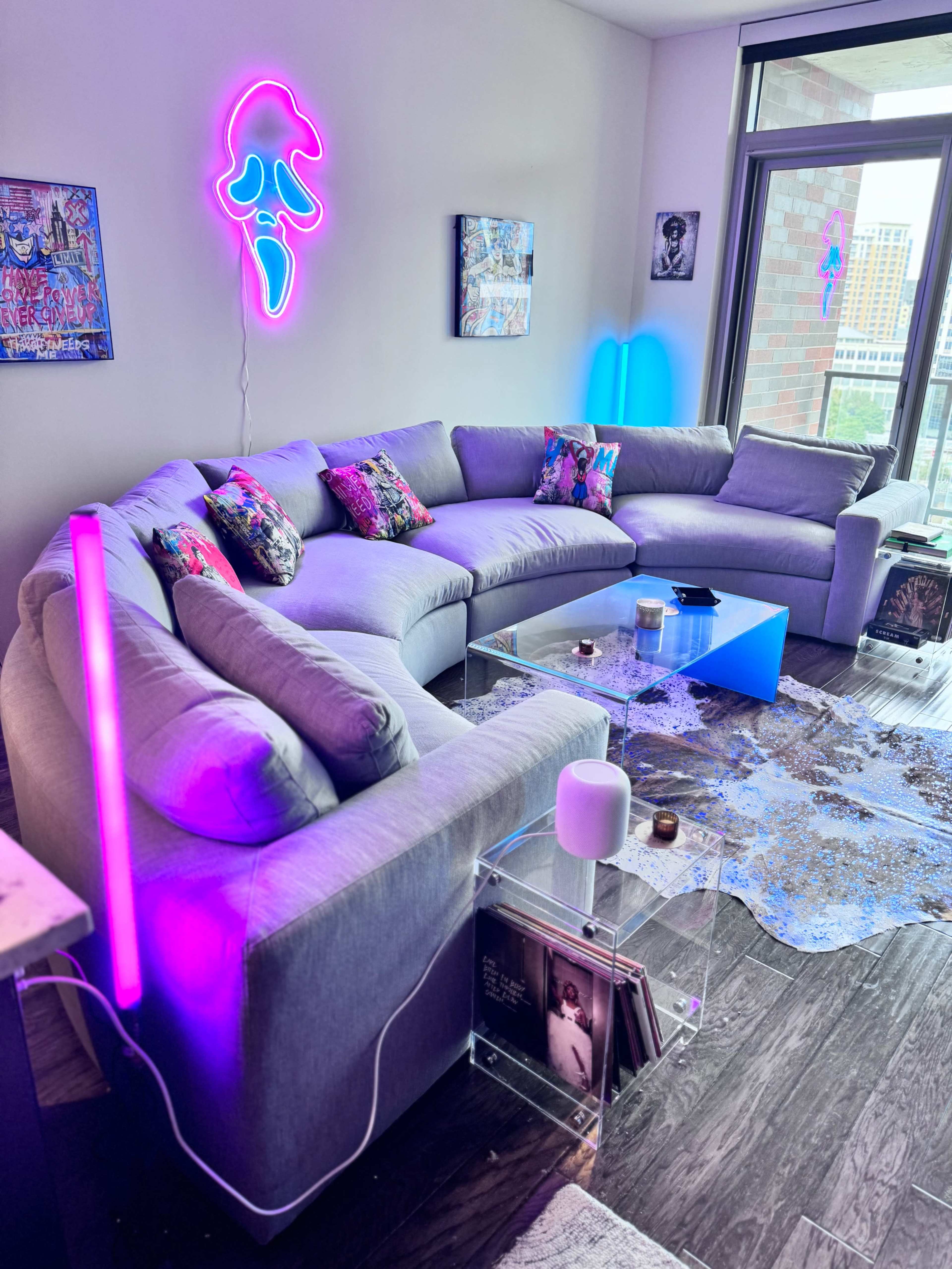 A modern living room with a gray sectional sofa adorned with colorful pillows, a transparent coffee table, and a neon light feature on the wall.