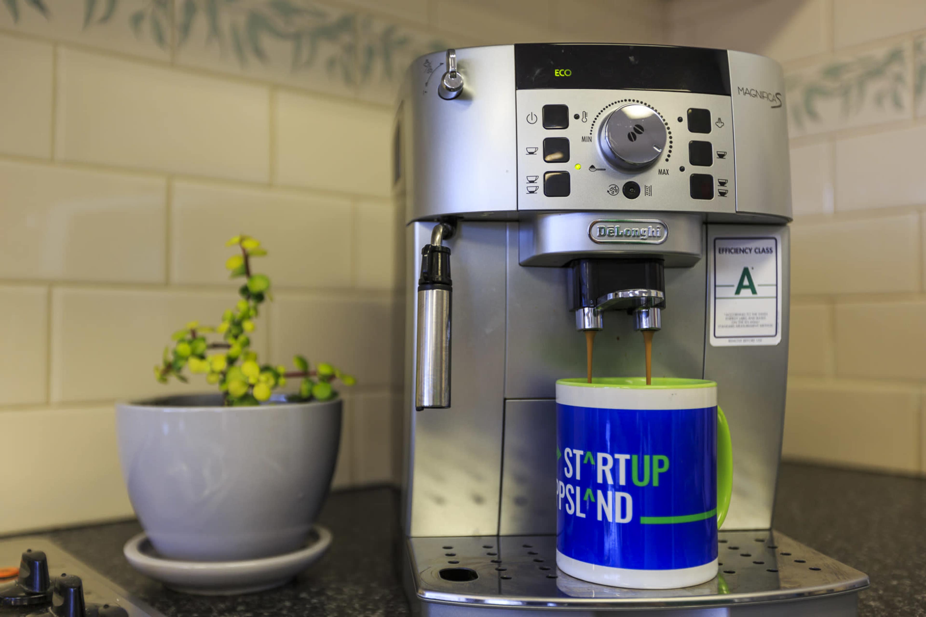 A coffee machine is brewing a cup of coffee into a blue and green mug labeled "STARTUP."