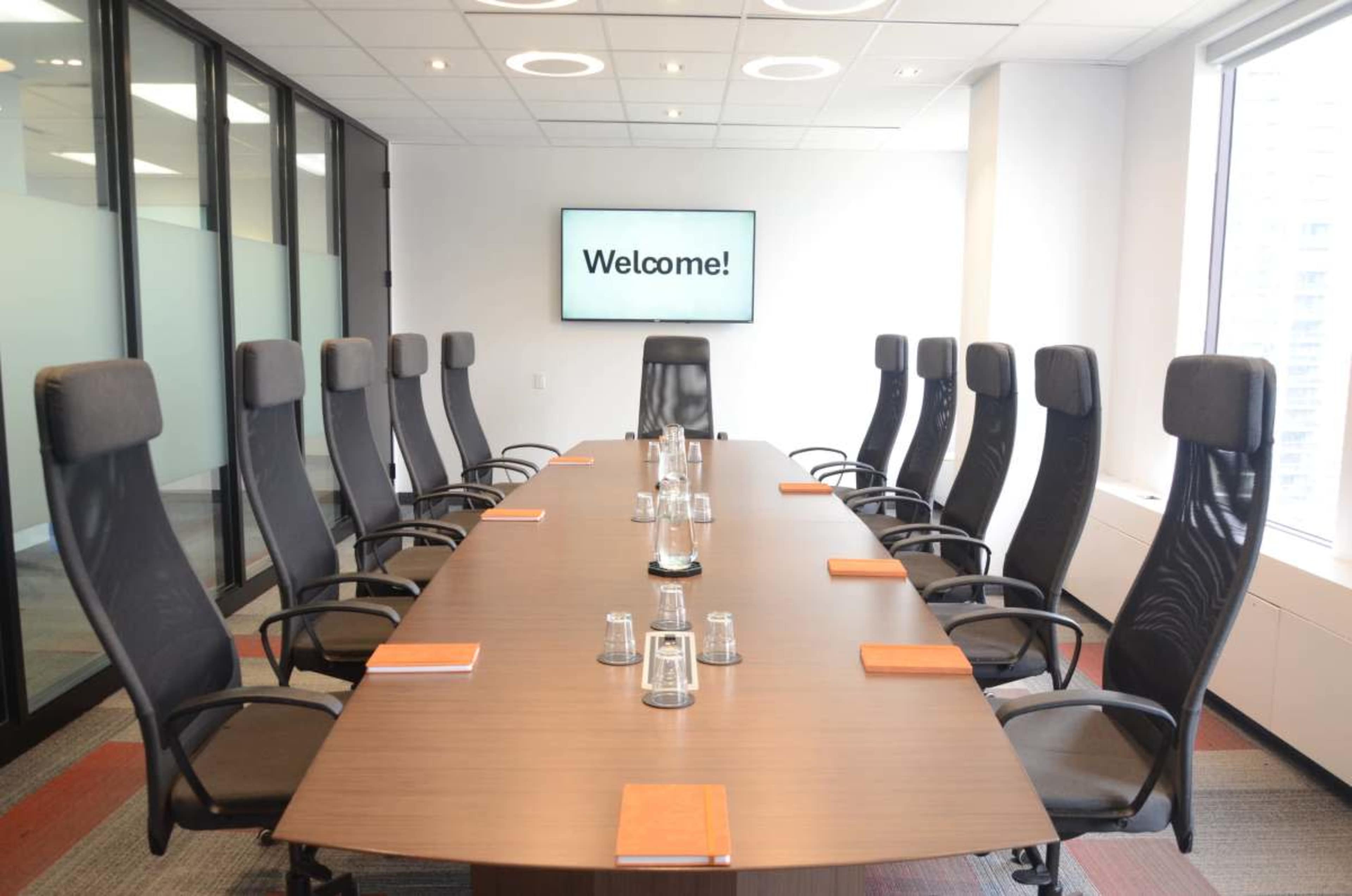 A well-lit conference room features a large table surrounded by eight high-backed chairs, with a screen displaying the message "Welcome!" at one end.