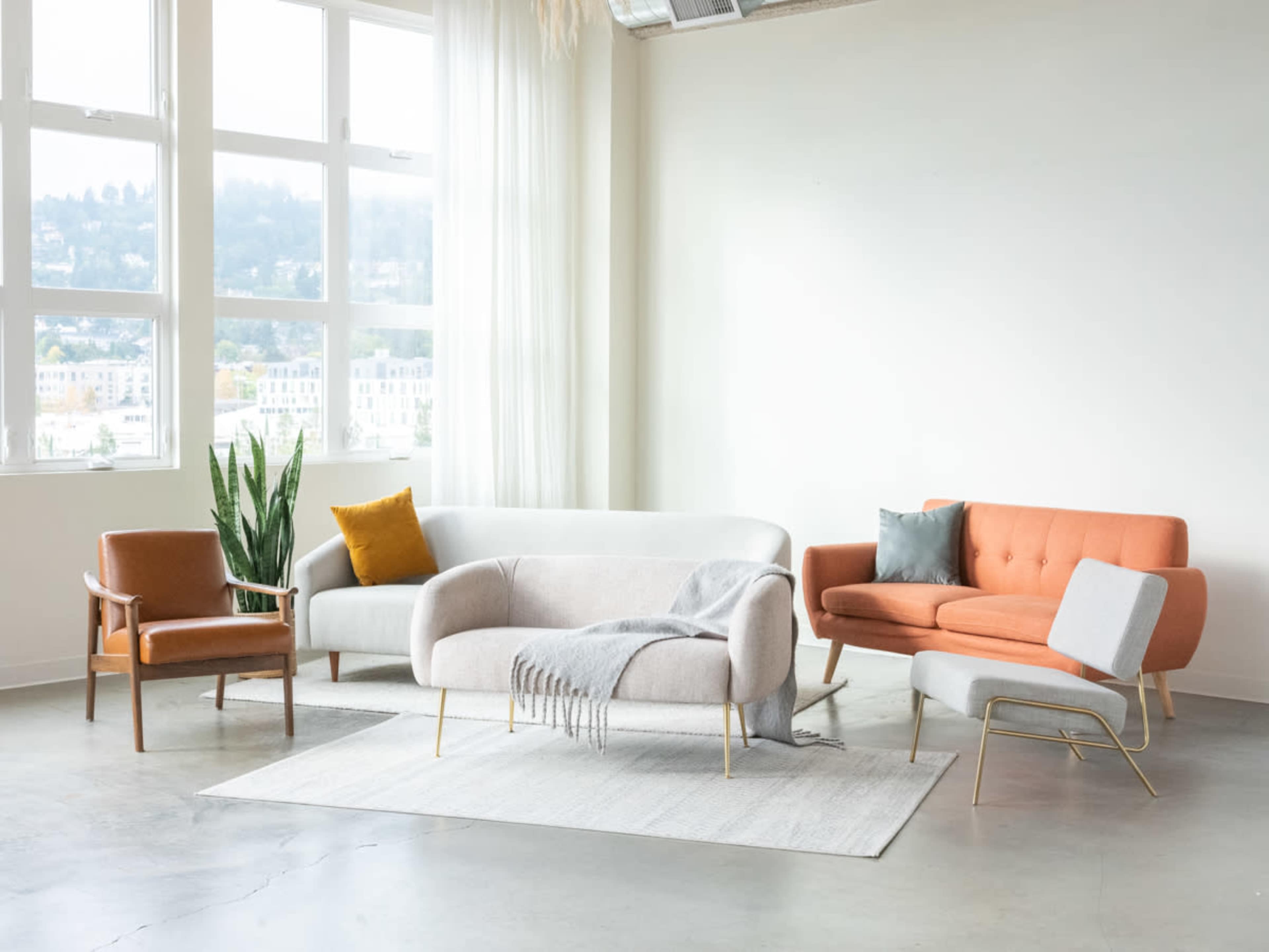 The image shows a modern living room setup featuring a gray and orange sofa, a light gray loveseat, a small chair, and a decorative plant, all arranged on a light rug in a well-lit space with large windows.