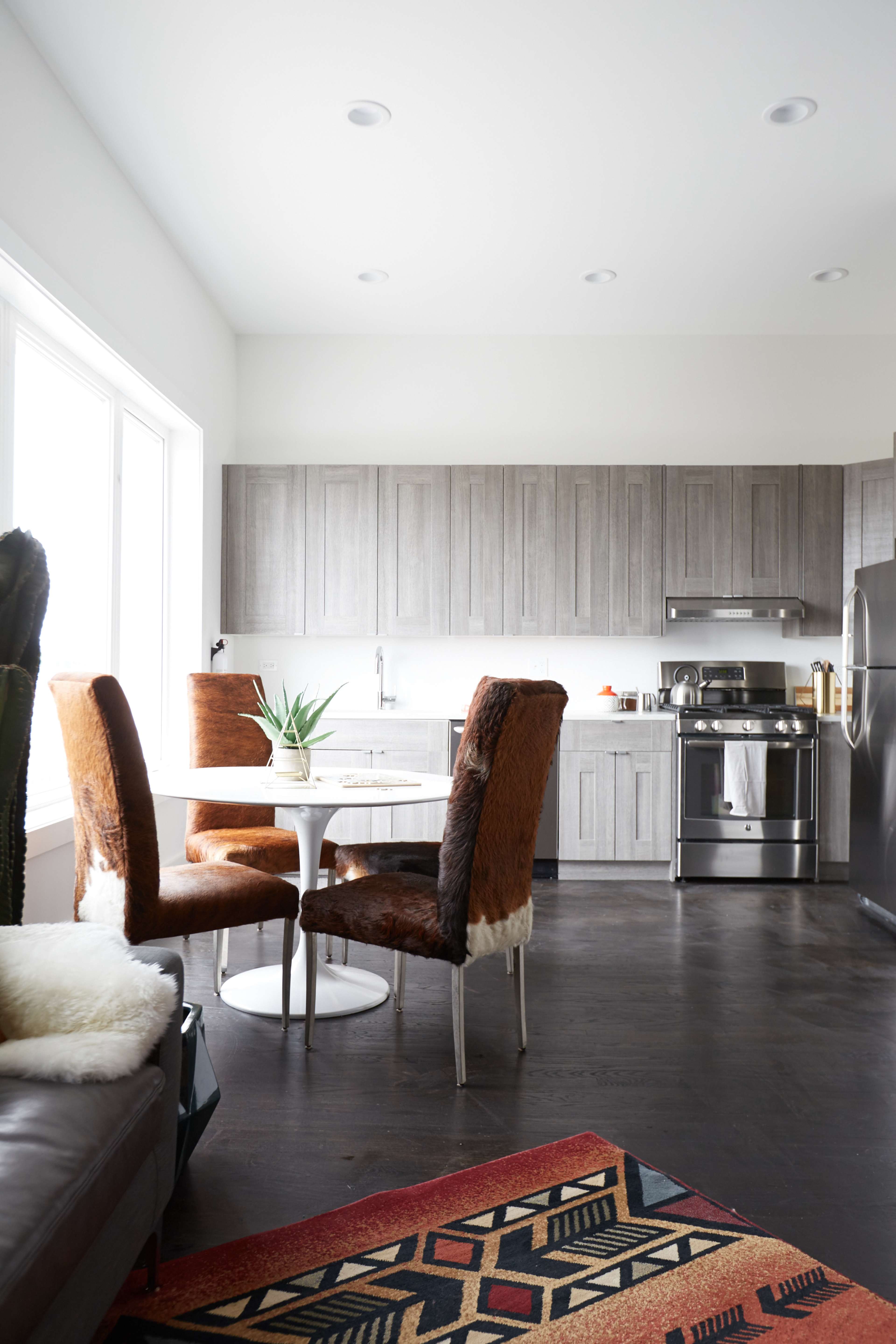 A modern kitchen features a round table with four chairs, gray cabinets, stainless steel appliances, and a patterned rug on a dark floor.