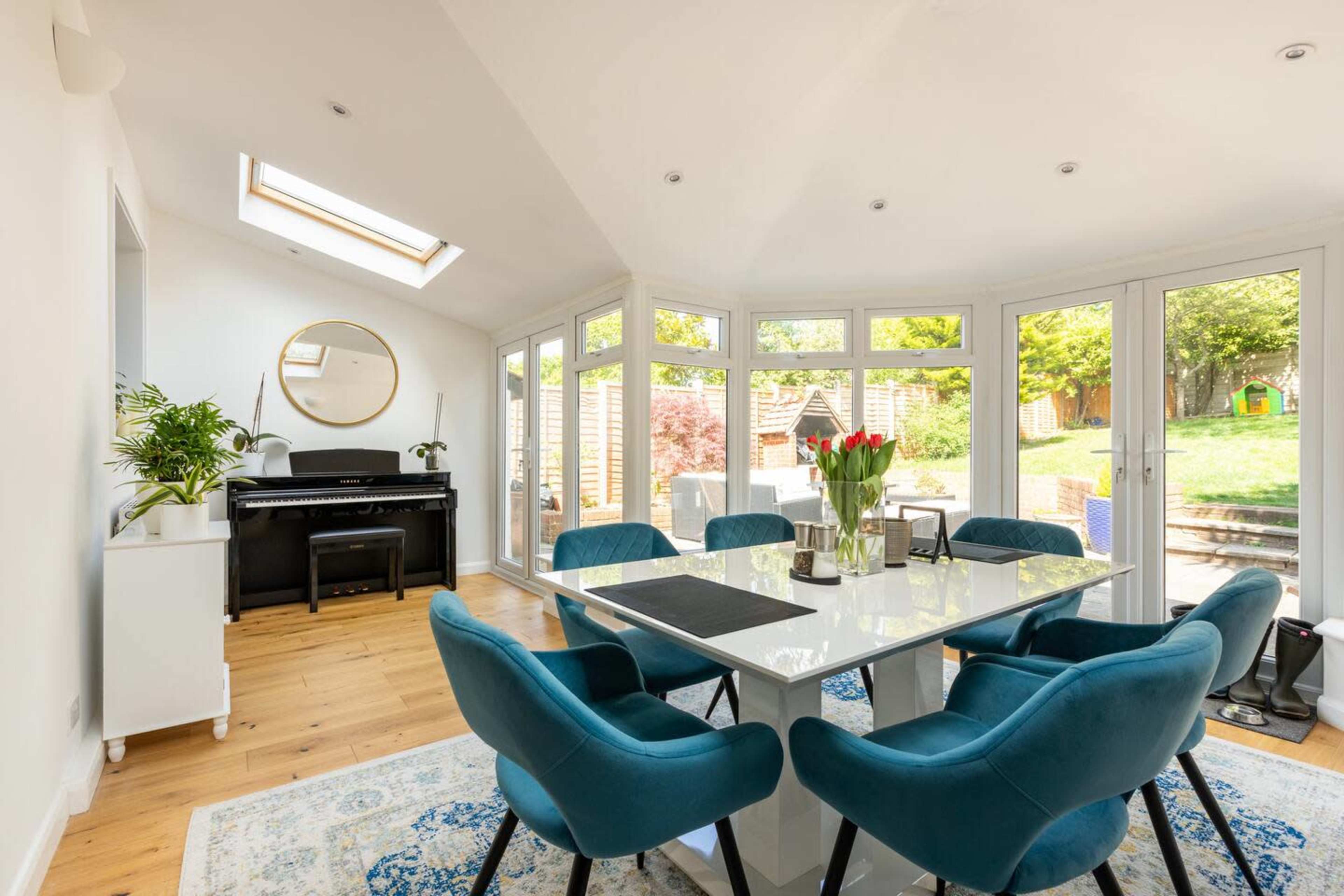 A bright dining area features a large table surrounded by blue chairs, with a piano and windows that overlook a garden.
