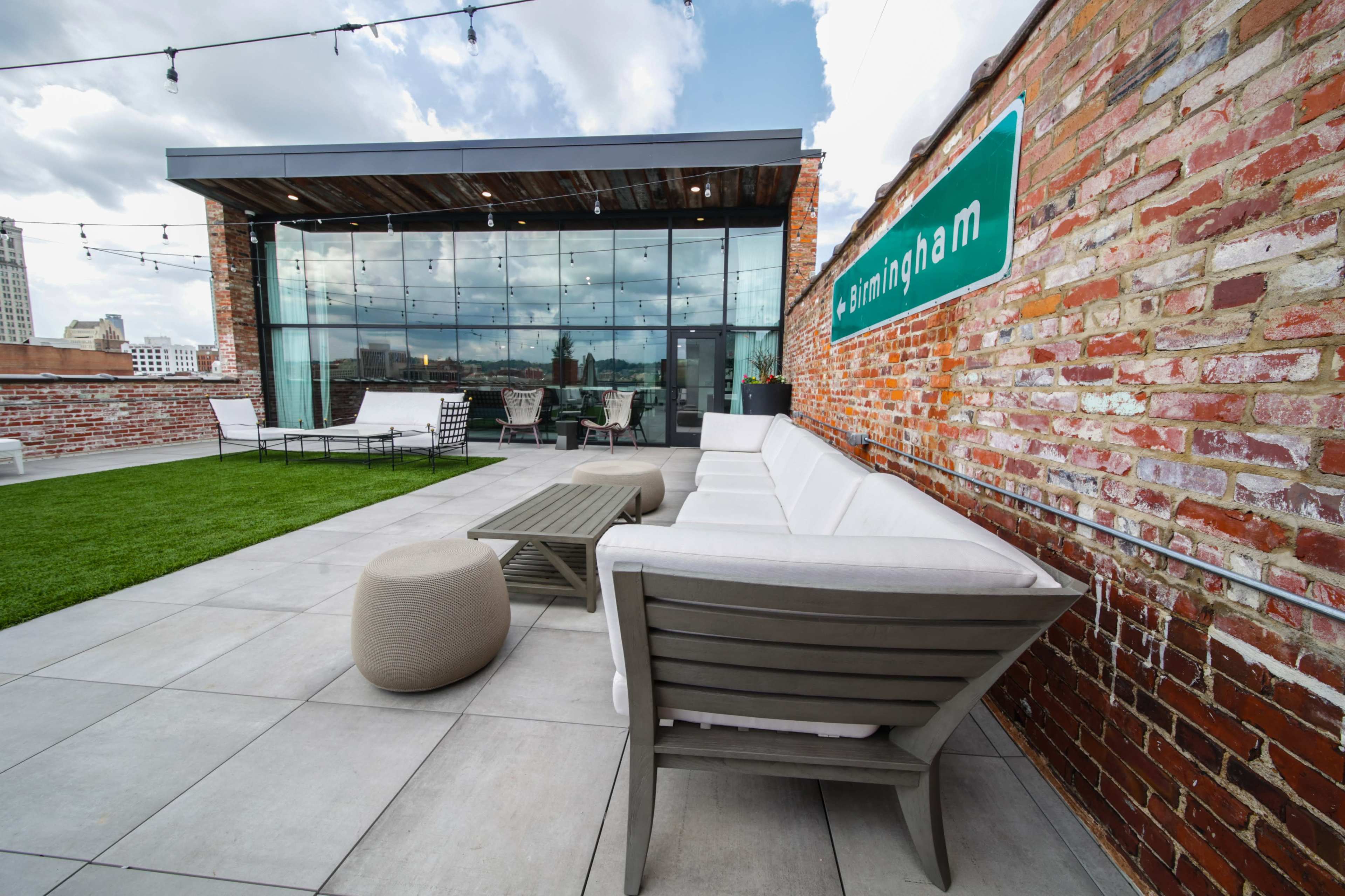 The image shows a modern rooftop terrace with comfortable seating, a grassy area, and large glass windows overlooking a cityscape.