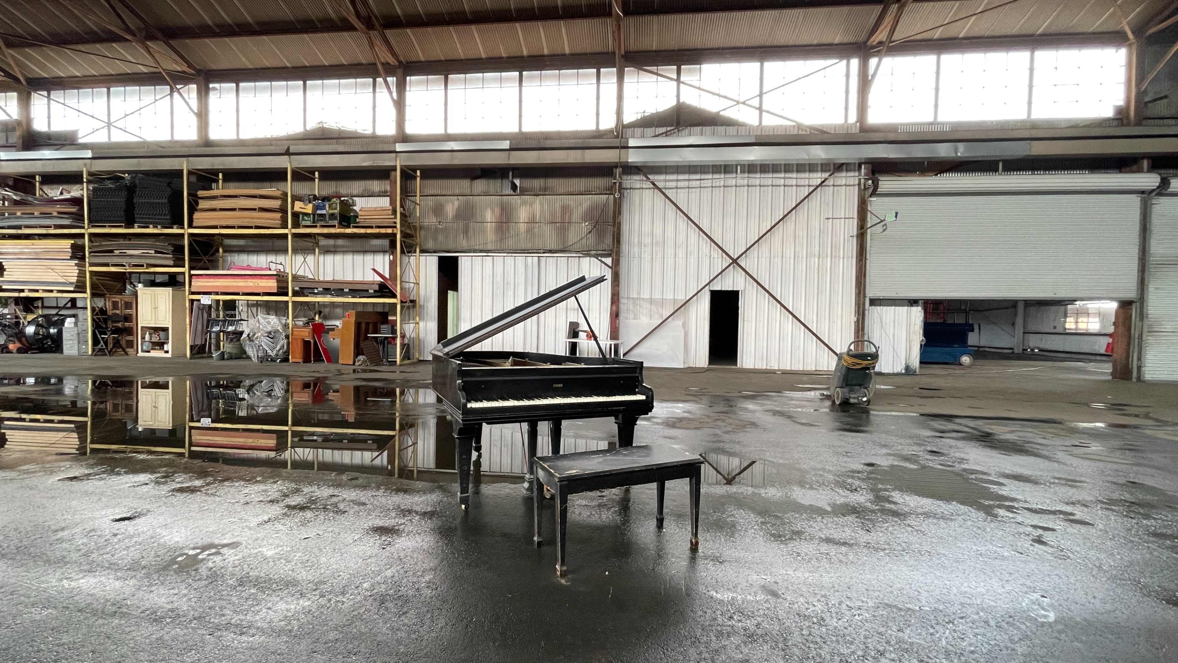 A grand piano and its bench sit on a wet, reflective floor inside an industrial warehouse with large open doors.