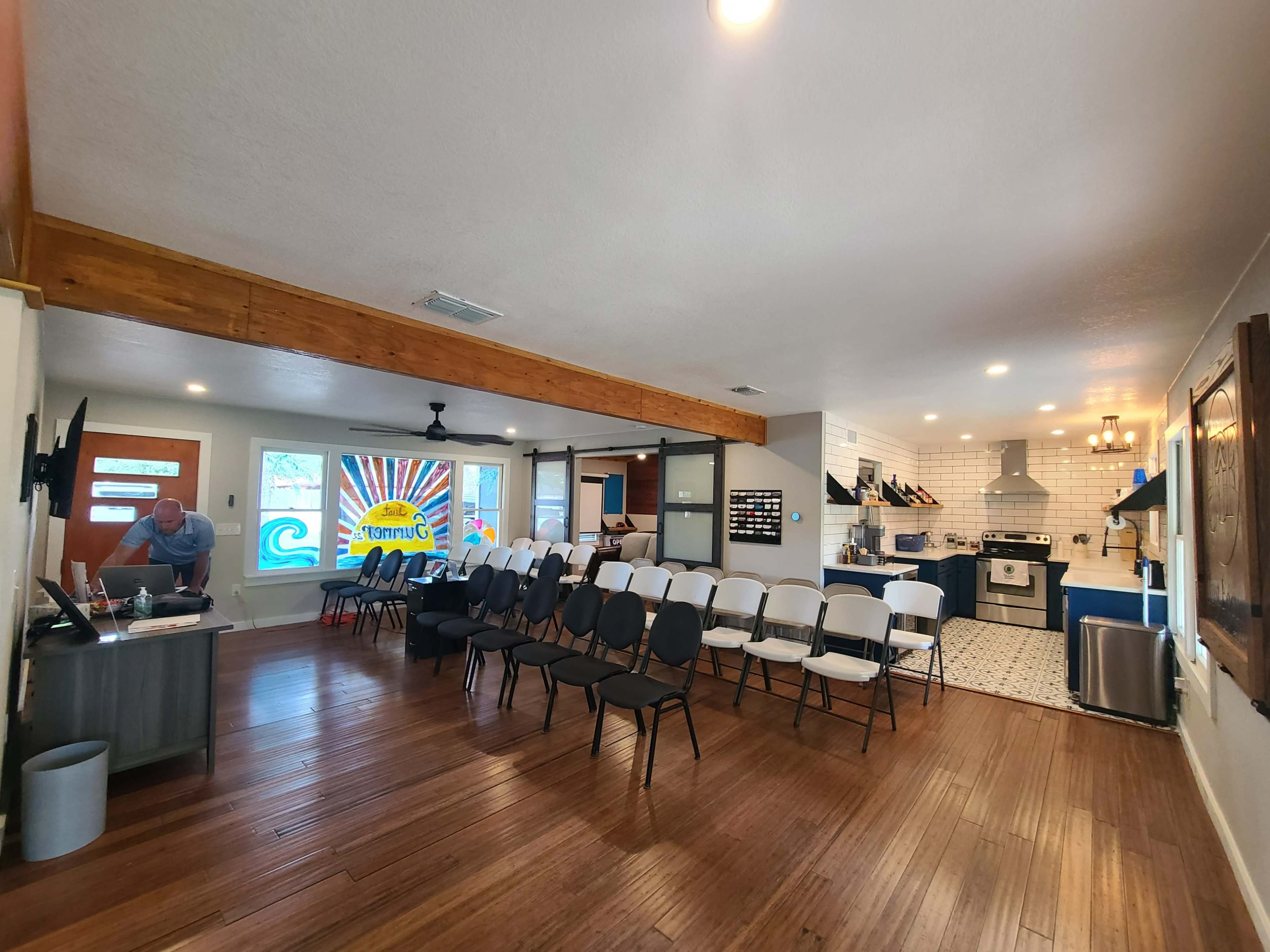 The image shows a spacious room with wooden flooring, featuring rows of chairs, a modern kitchen area, and colorful wall decor.