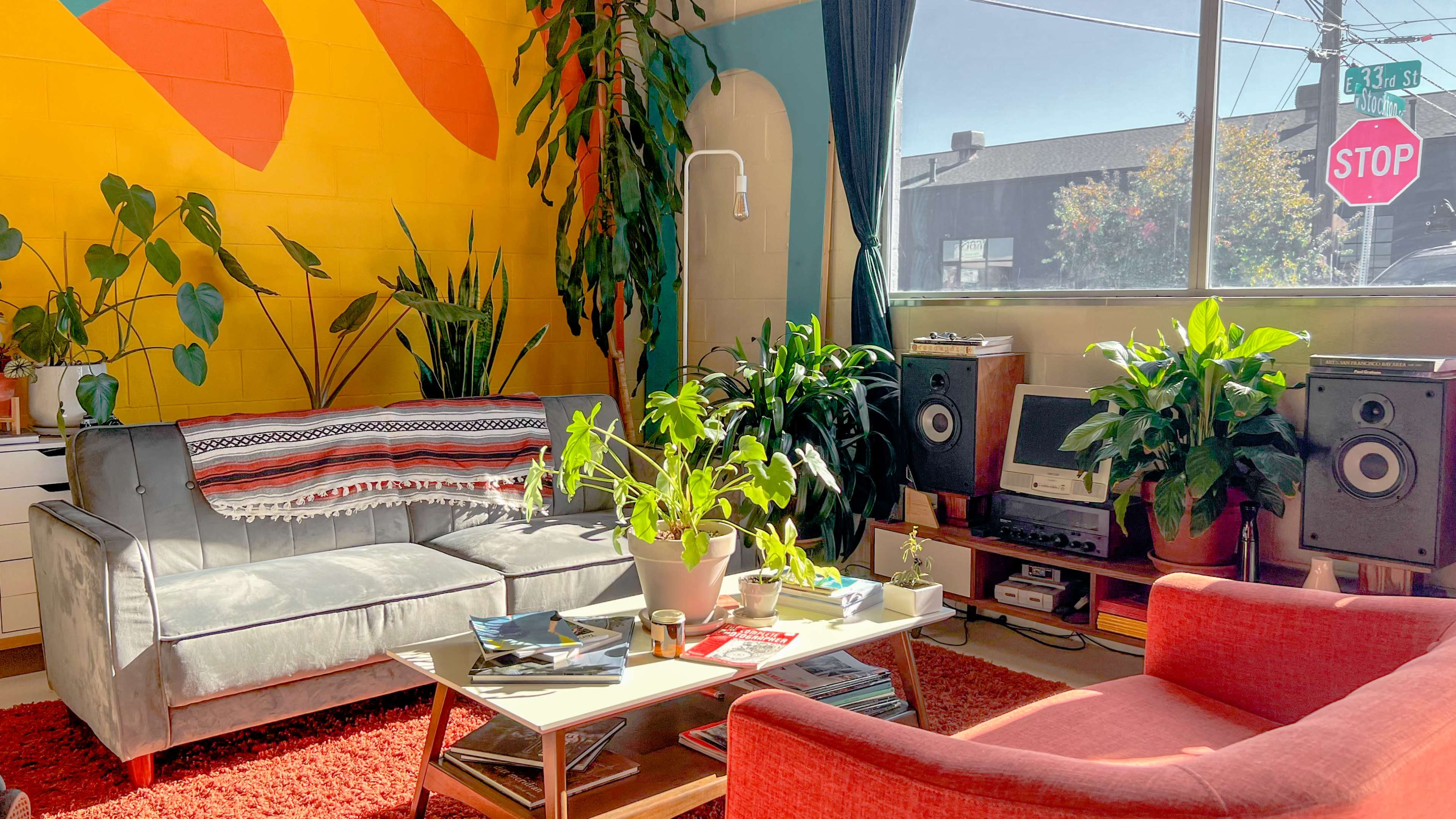 The scene features a brightly colored living room with plants, a gray sofa, a red armchair, a coffee table, and audio equipment against a vibrant mural.