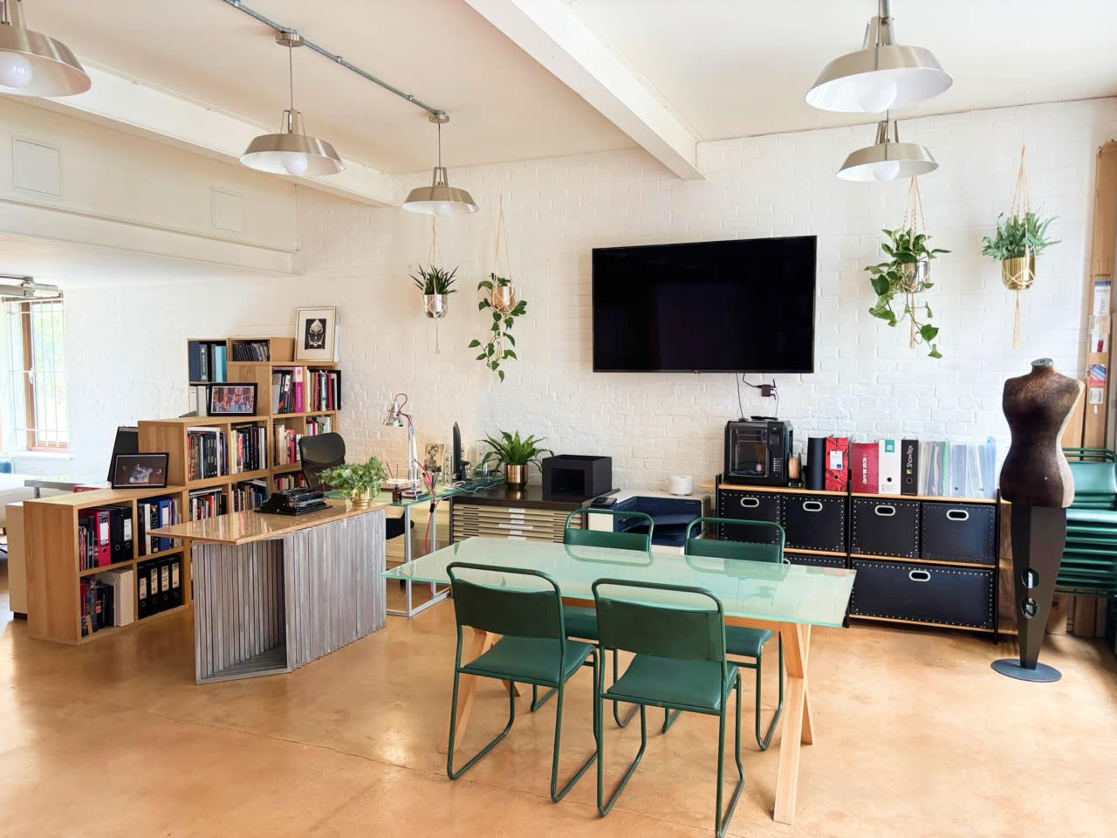 The image depicts a modern office space featuring a large table with green chairs, a bookshelf filled with books, a television mounted on a white wall, and various potted plants.
