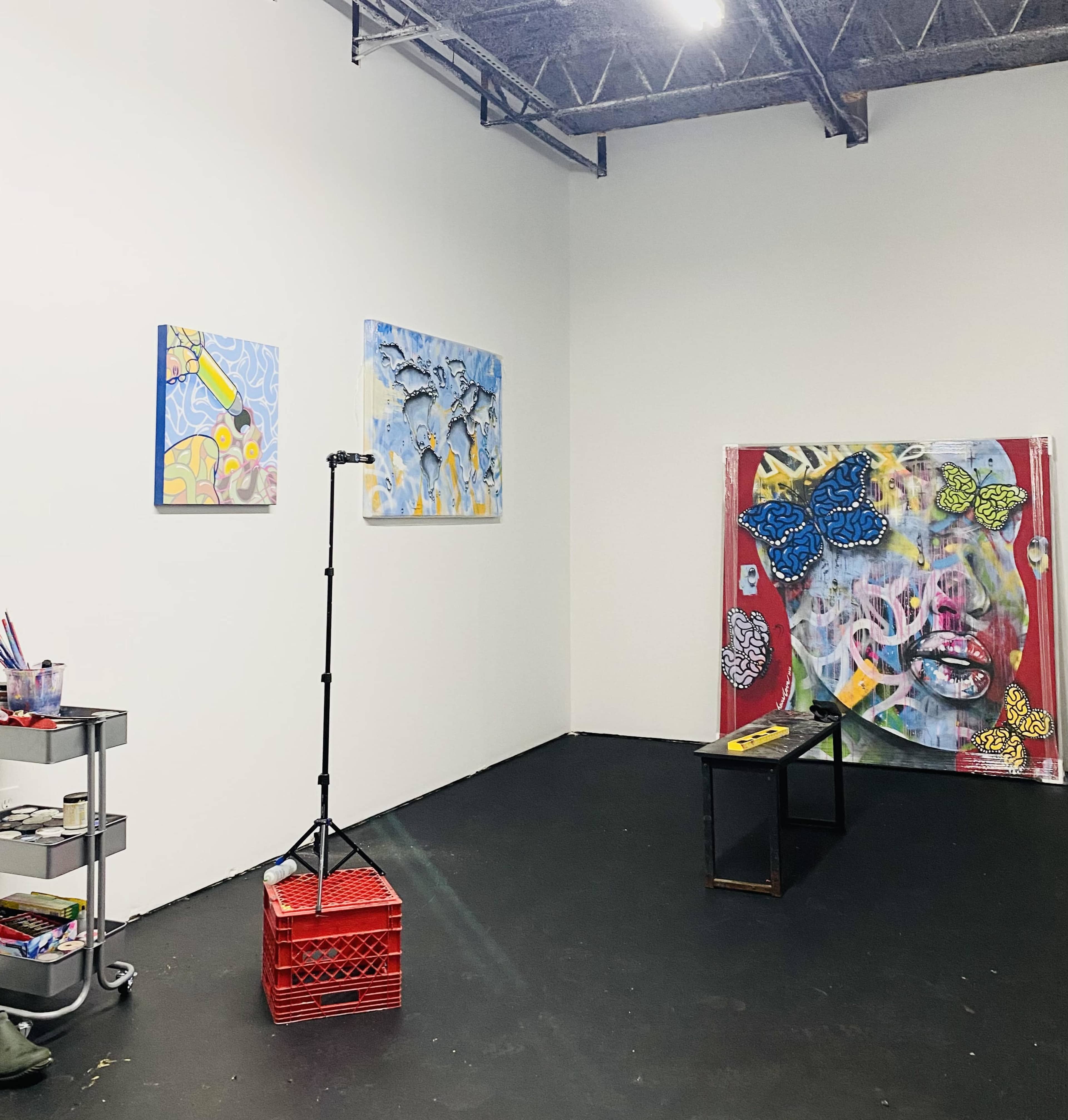An art studio with two abstract paintings on the wall, a large colorful artwork, and a cart with art supplies in one corner.