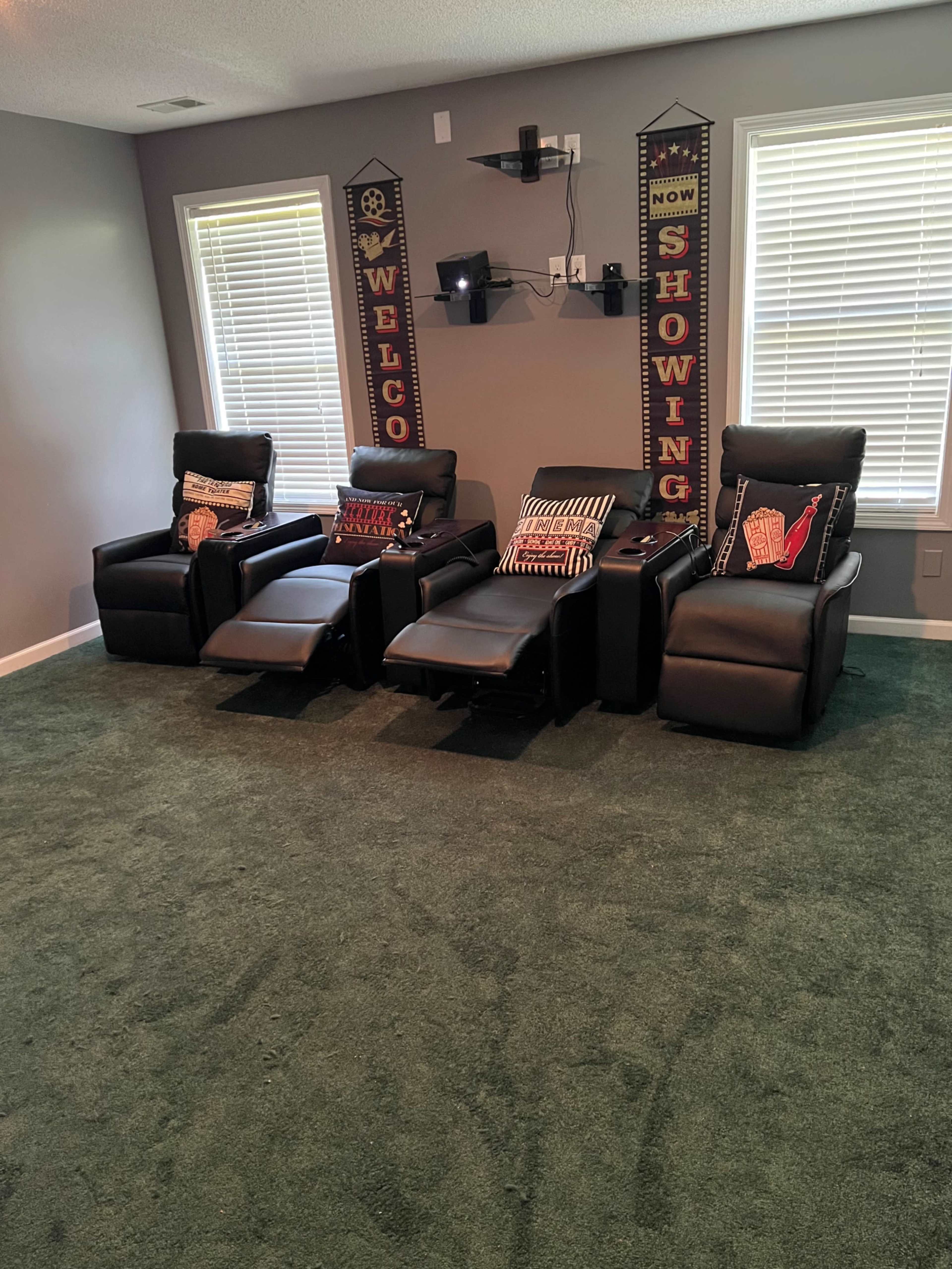 The image shows a home theater setup featuring three black recliner chairs positioned in front of a wall with dual decorative banners and a mounted shelf.