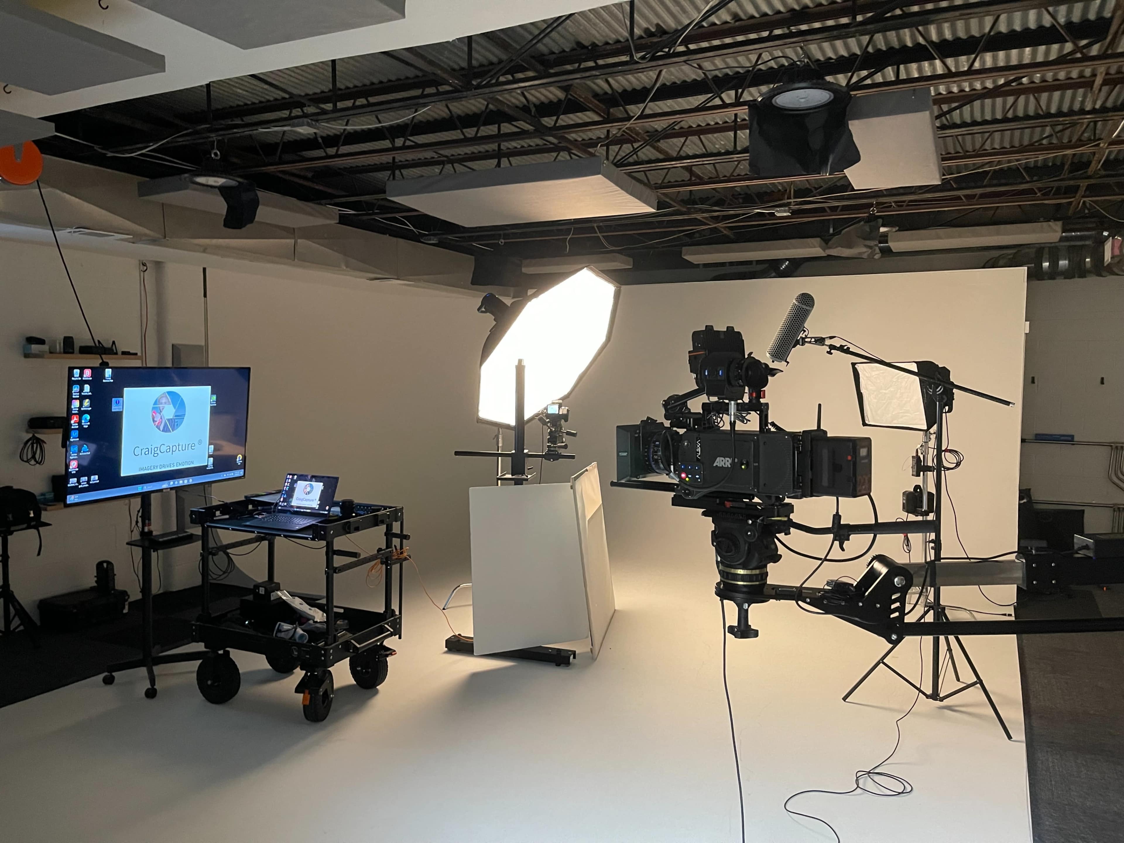 A video production setup features a camera on a tripod, an illuminated backdrop, a computer display on a cart, and various lighting equipment in a studio environment.