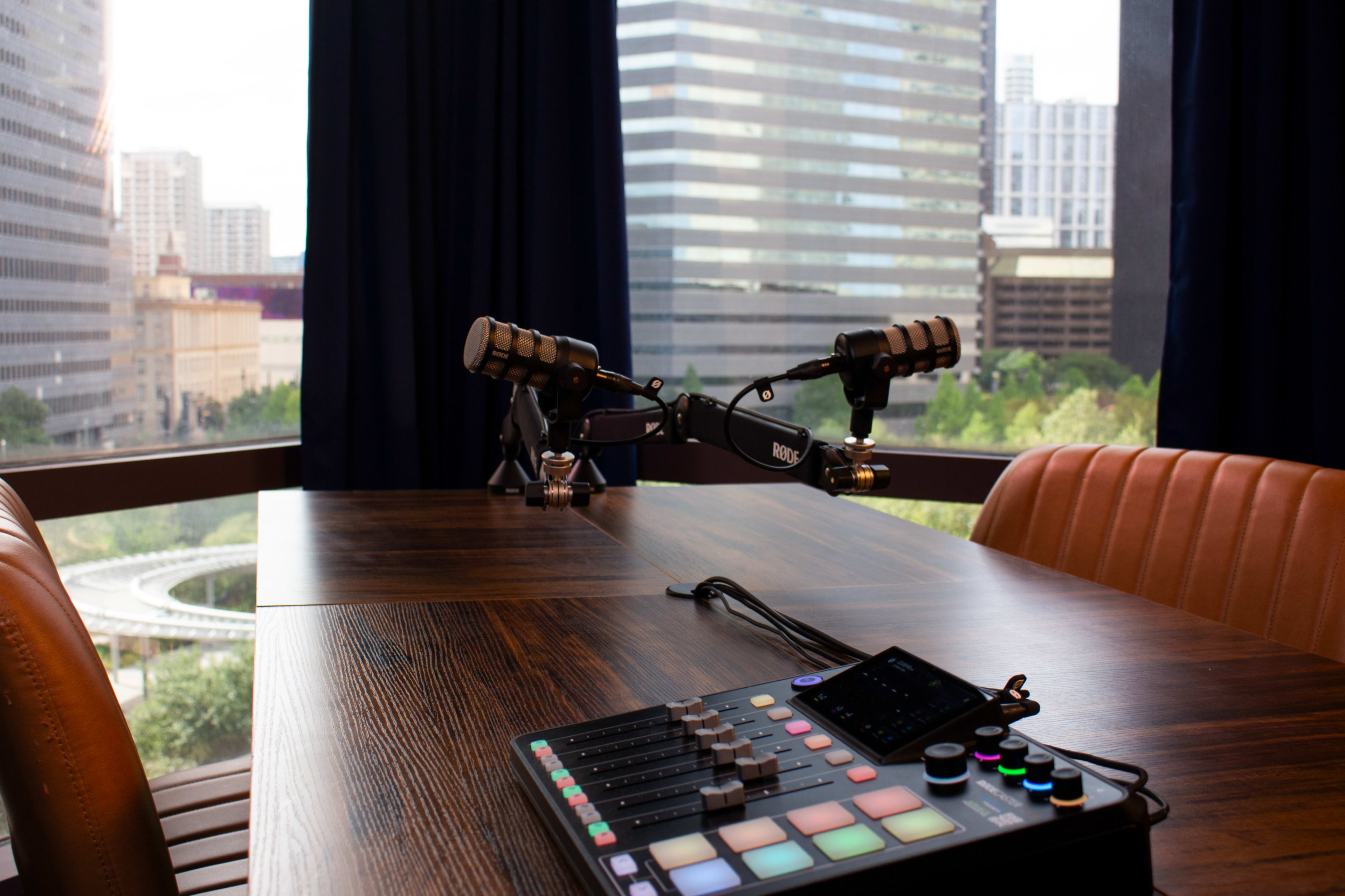 A modern podcasting setup featuring two microphones and a soundboard on a wooden table, with a view of a city skyline through large windows.