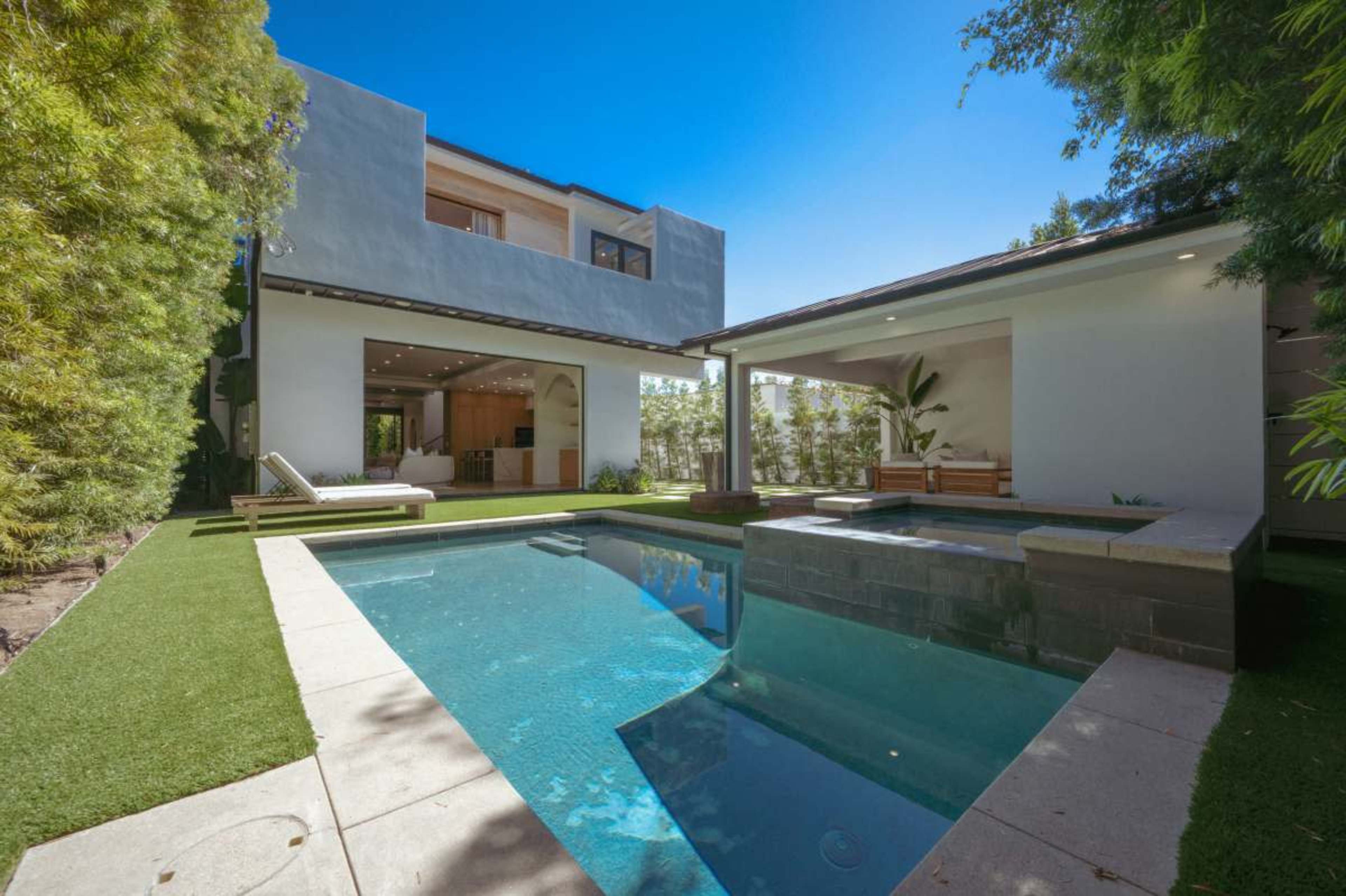 The image shows a modern outdoor space featuring a swimming pool, landscaped greenery, and a house with large windows in the background.