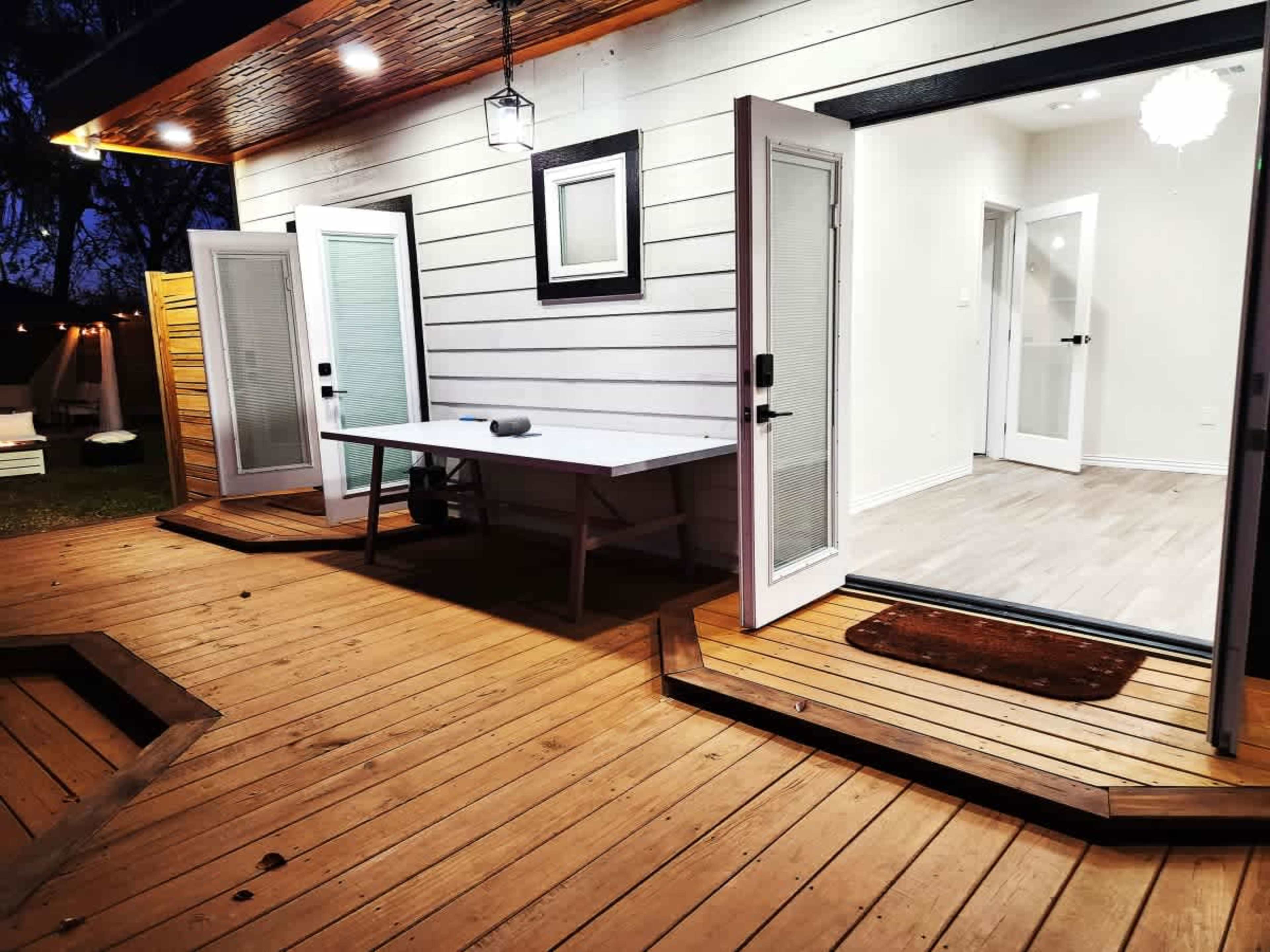 The image shows a modern outdoor deck with wooden flooring, featuring two sets of double doors that lead into an interior space.