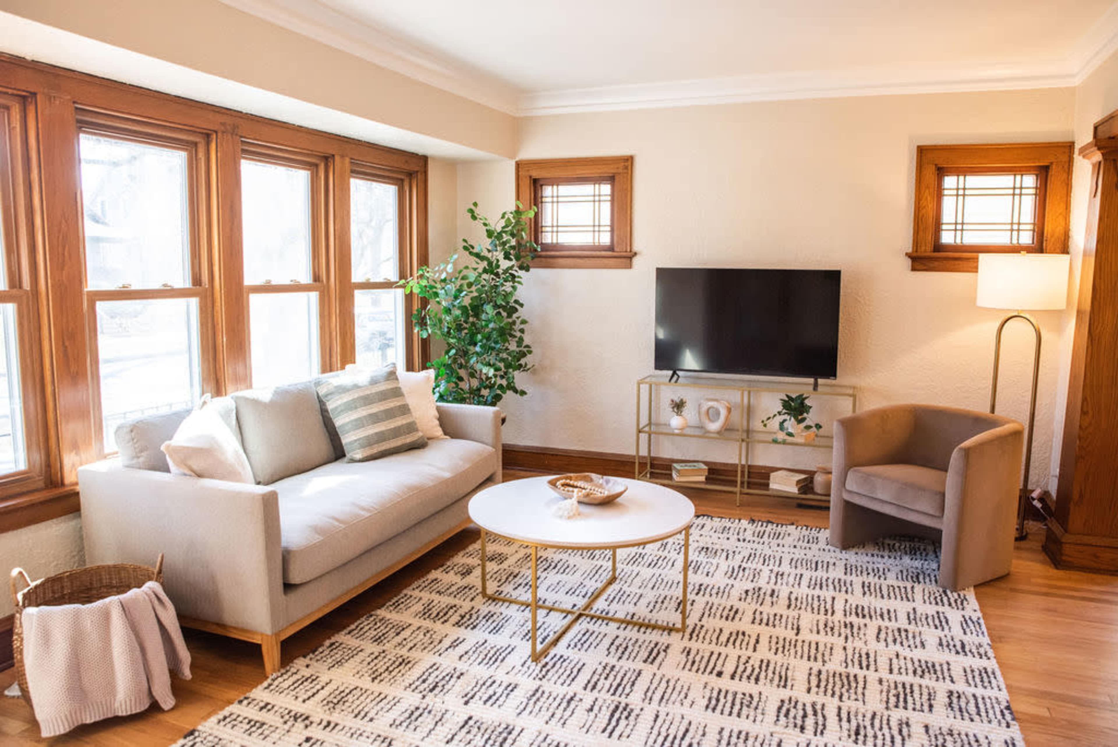 A well-lit living room featuring a beige sofa, a round coffee table, a light-colored armchair, a television mounted on the wall, and multiple windows.