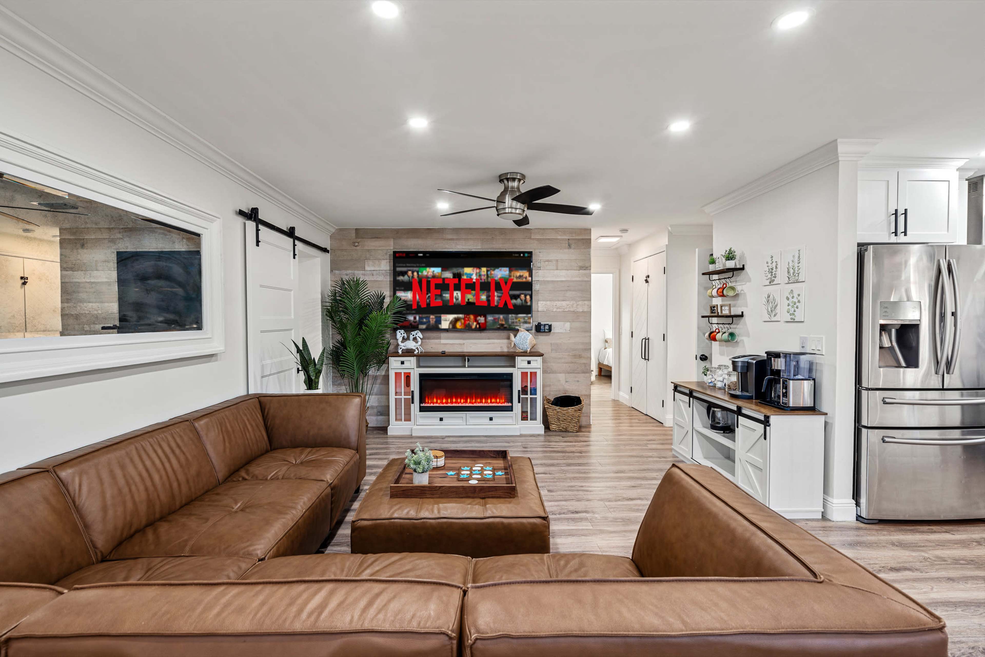A modern living room features a brown sectional sofa, a wall-mounted television displaying the Netflix logo, and a sleek kitchenette in the background.