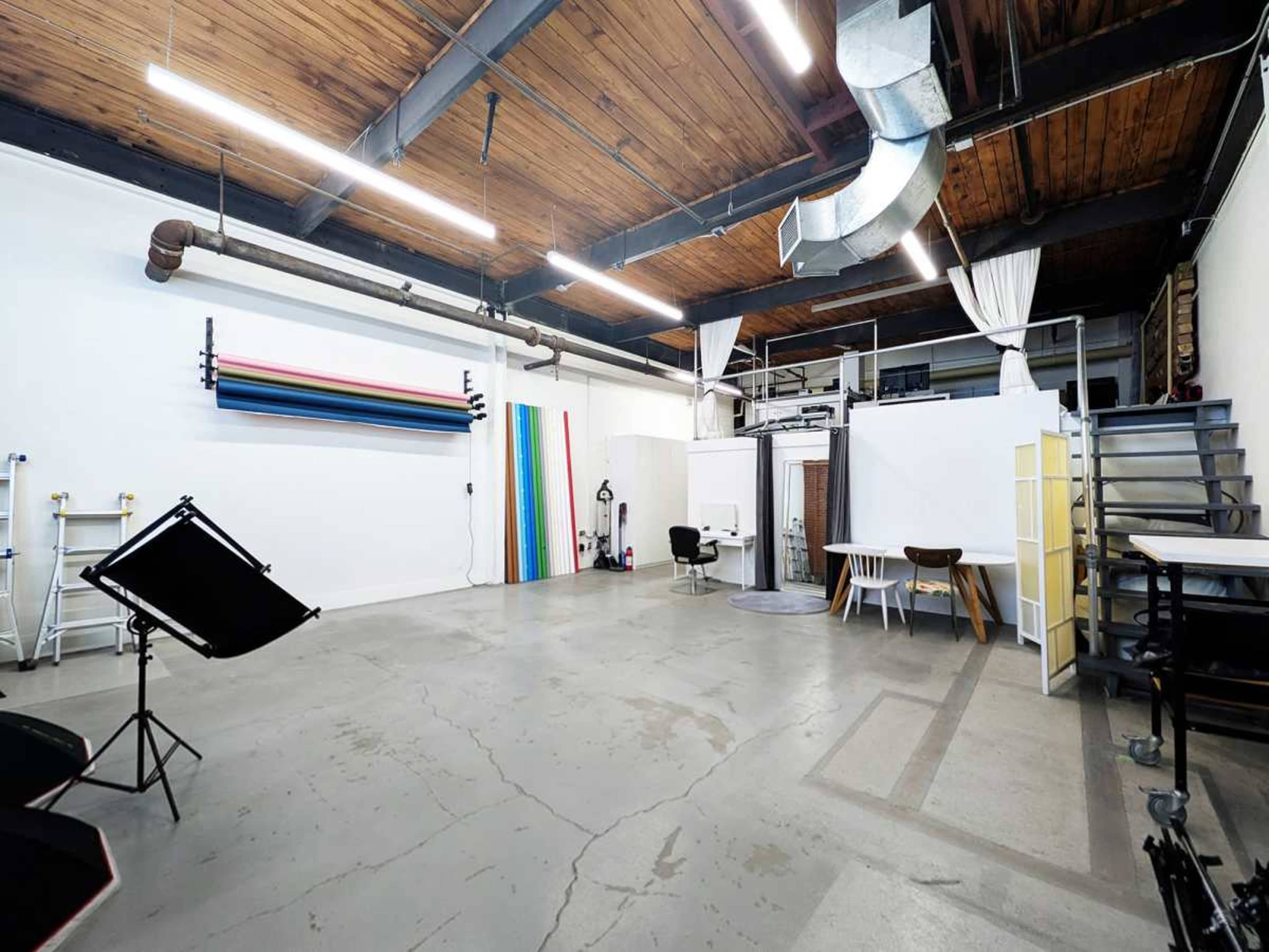 The image shows a spacious studio with high ceilings, featuring a variety of backdrops along one wall, and furniture arranged for a creative workspace.