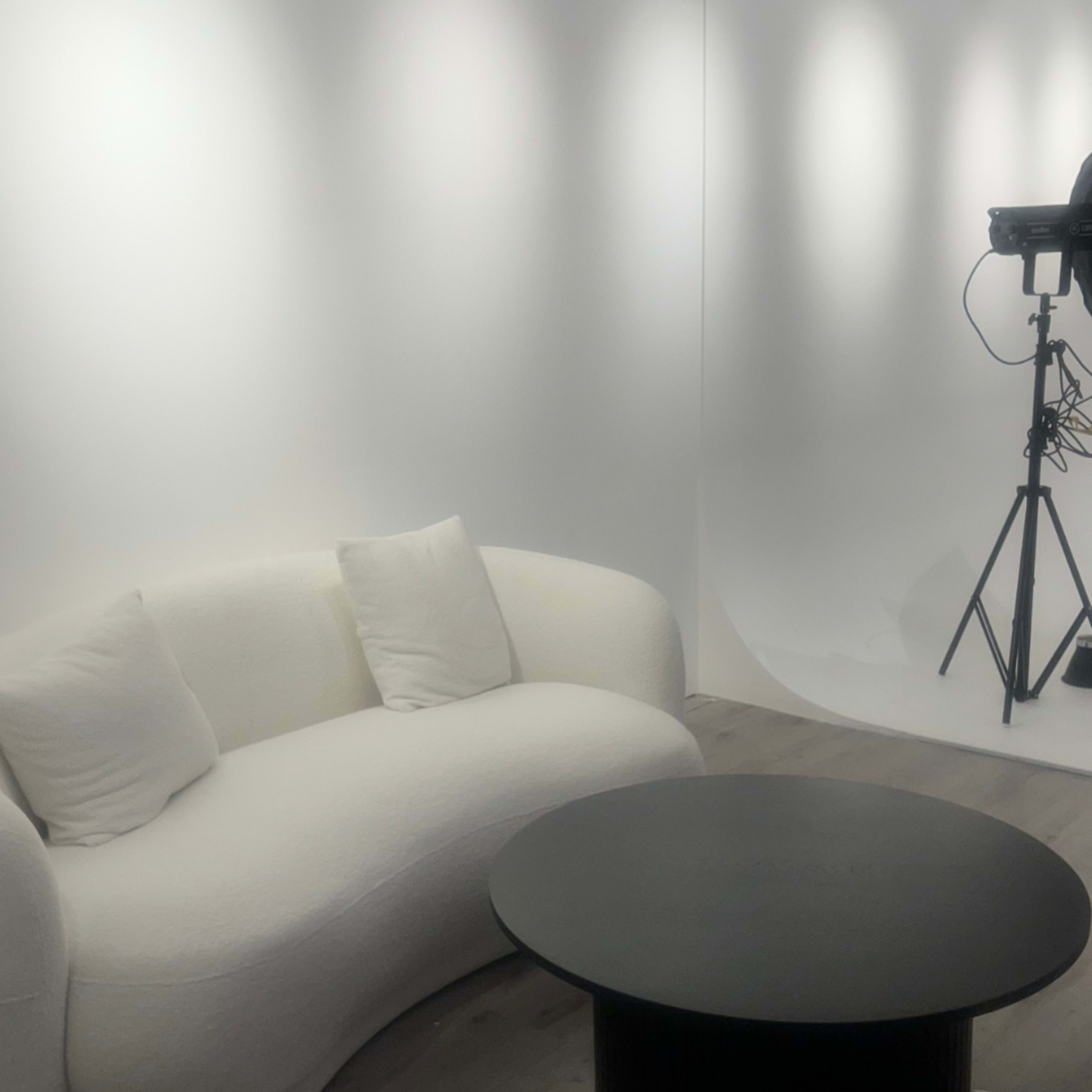 The image shows a minimalist studio space featuring a curved white sofa and a round black table, with studio lighting equipment in the background.