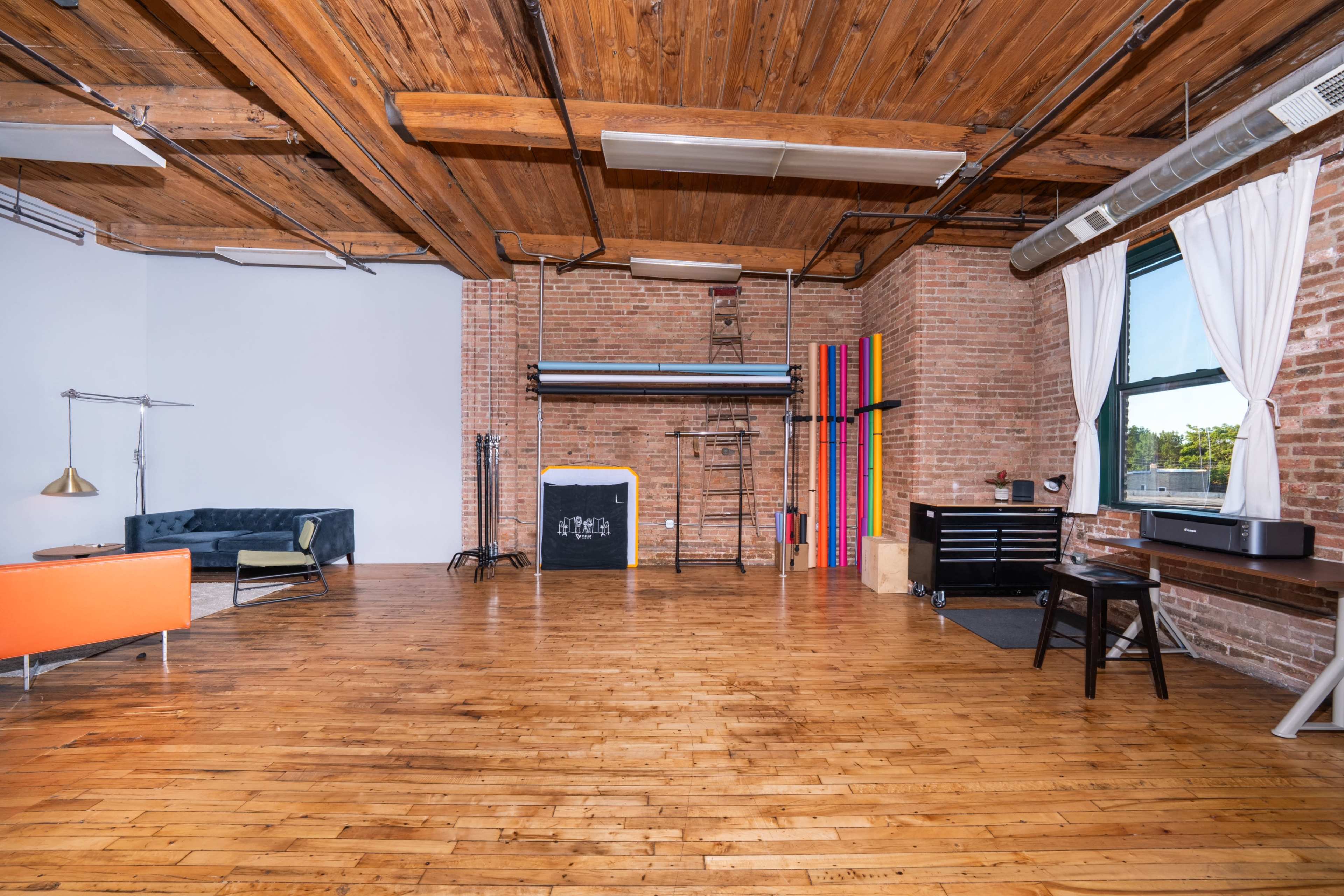 The image shows a spacious, industrial-style room with exposed brick walls, wooden flooring, and various furniture pieces, including a sofa and desk against one wall.