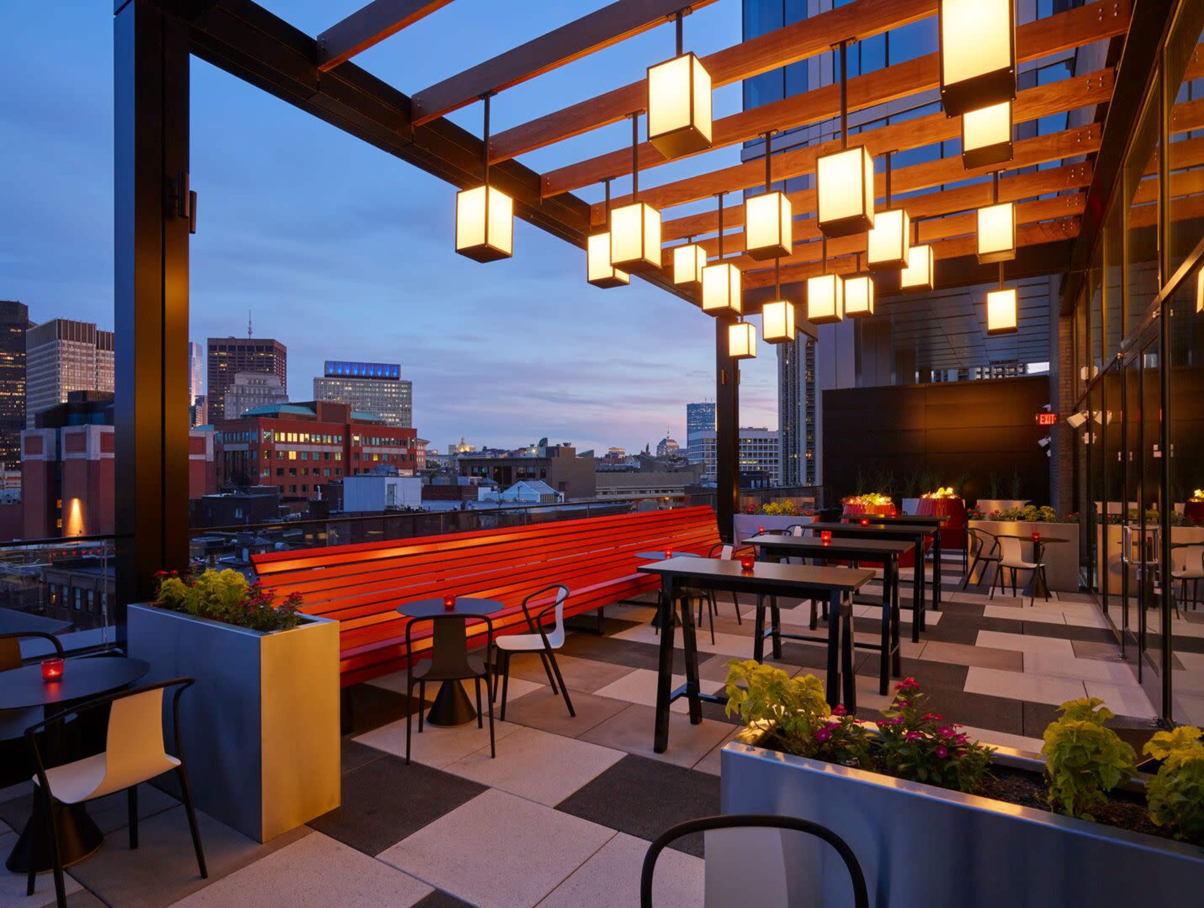 A modern rooftop dining area features wooden beams with hanging lanterns, surrounded by planters and a city skyline in the background during twilight.