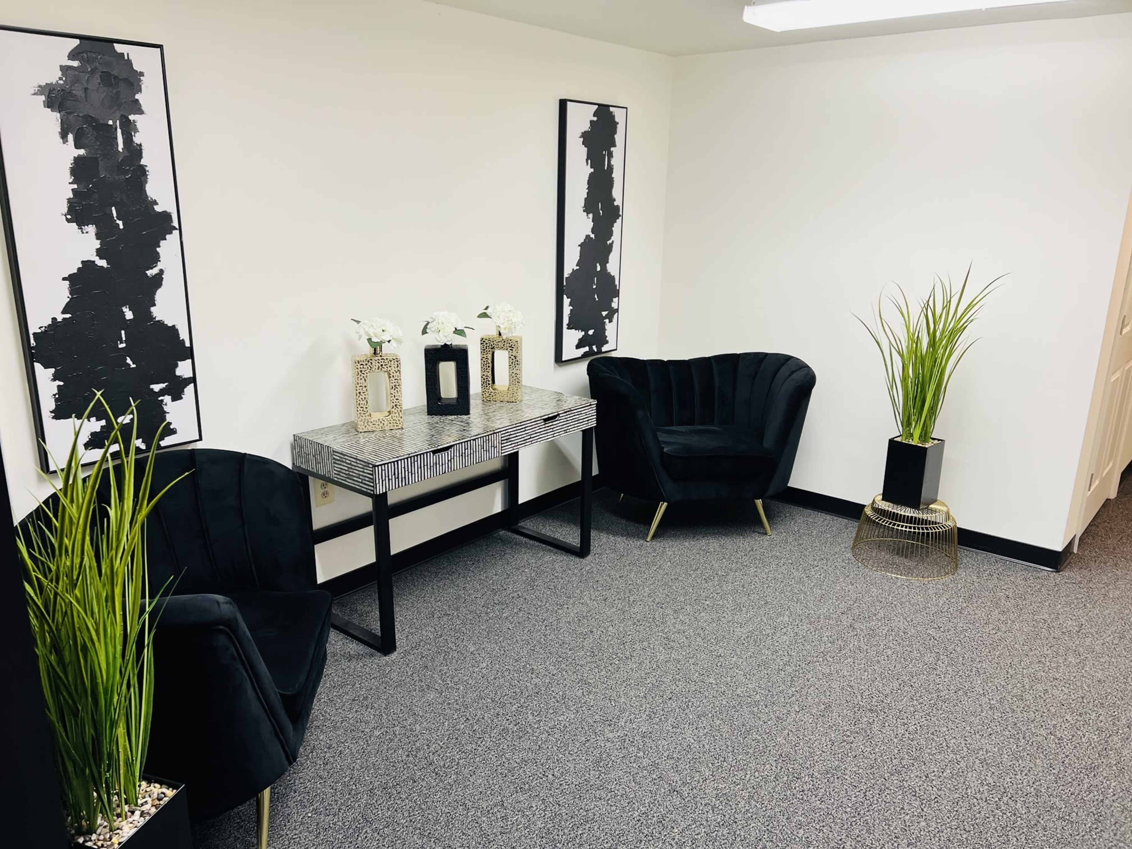 A minimalist office space with two black chairs, a patterned table, decorative plants, and framed artwork on the walls.