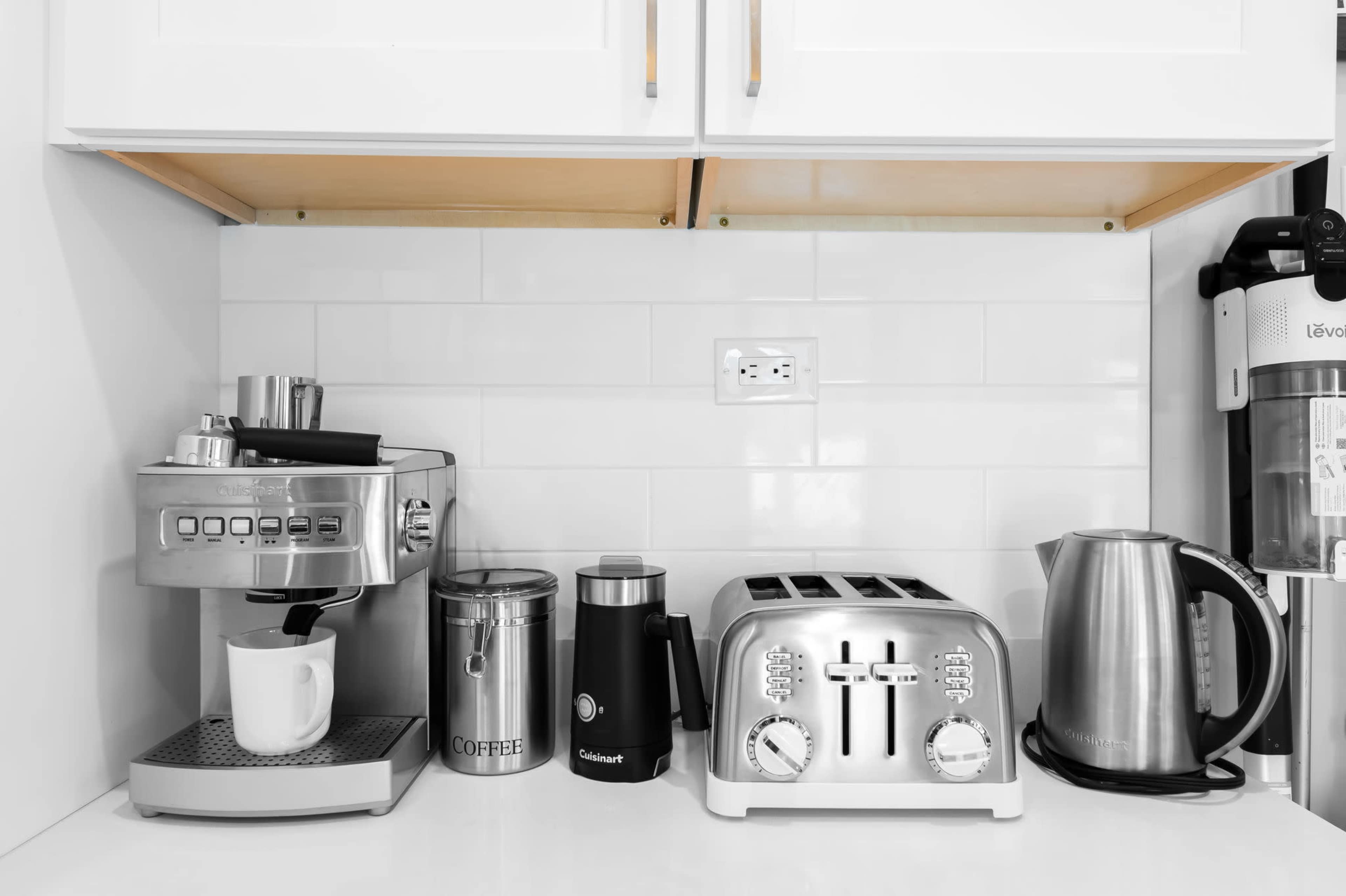 A coffee machine, toaster, kettle, and storage canister are neatly arranged on a countertop with white backsplash tiles.