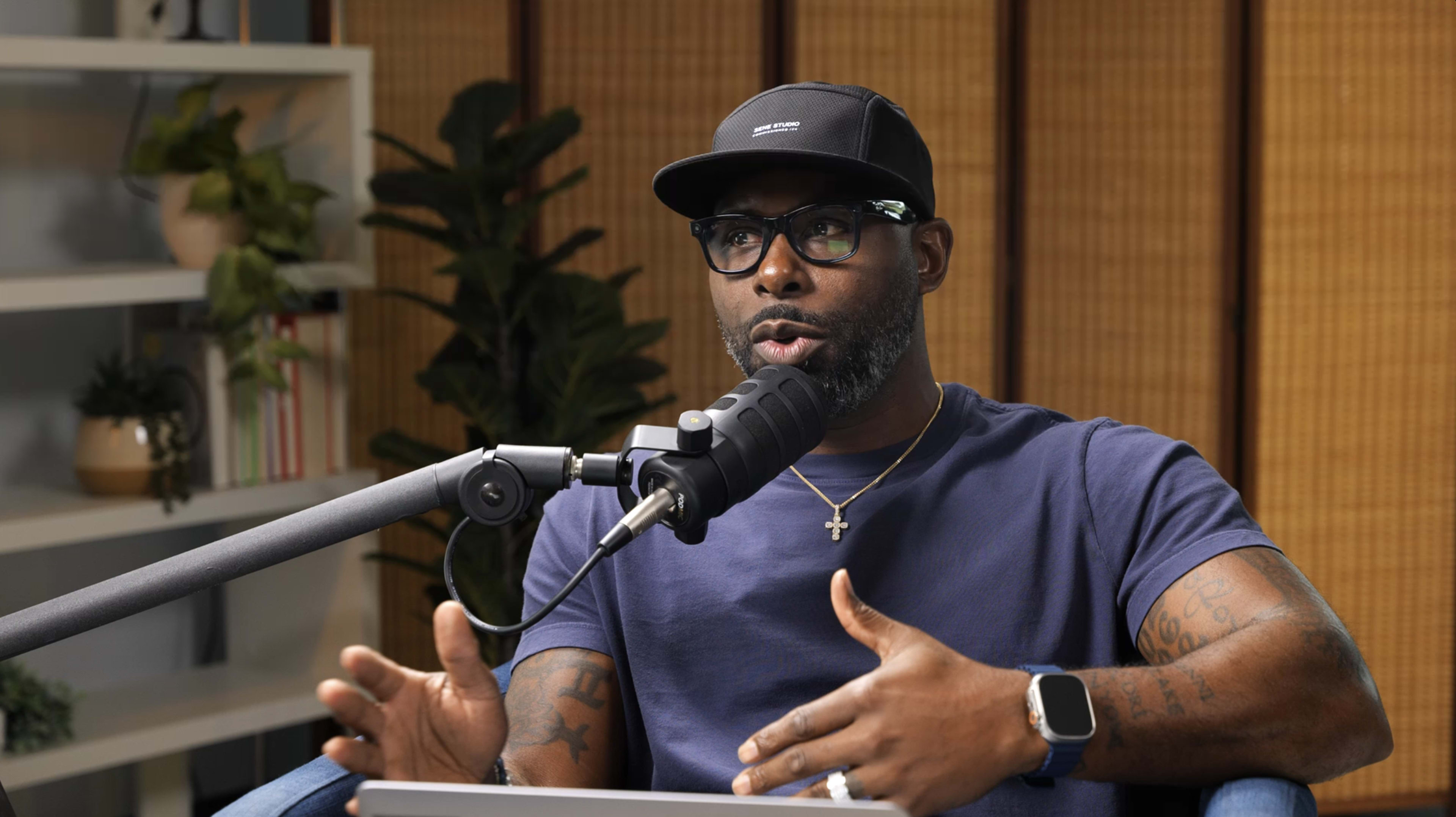 A man wearing glasses and a baseball cap is speaking into a microphone while sitting in front of a laptop in a well-lit room with plants in the background.