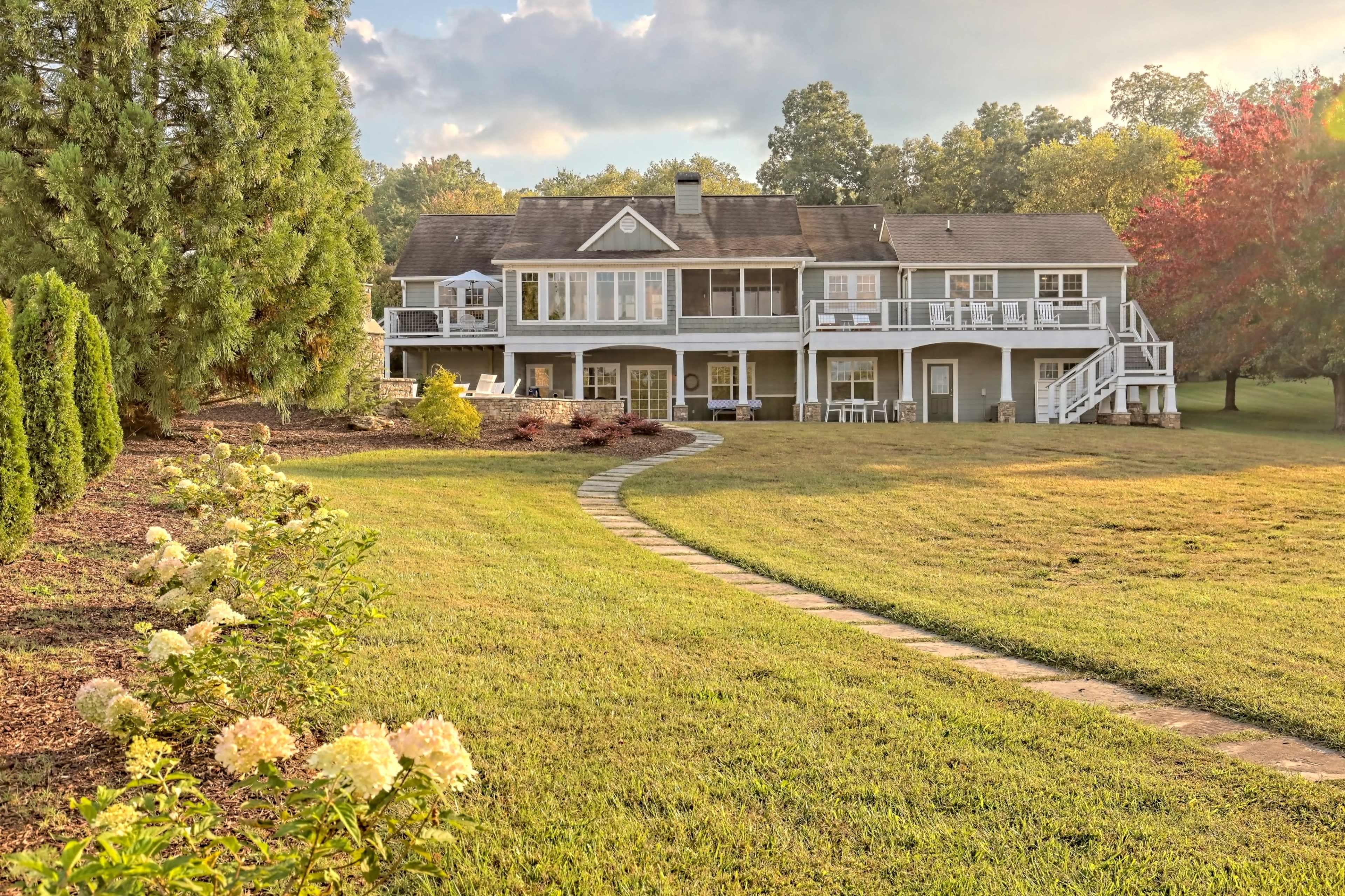 A two-story house with a large deck and a manicured lawn, surrounded by trees and a landscaped pathway.