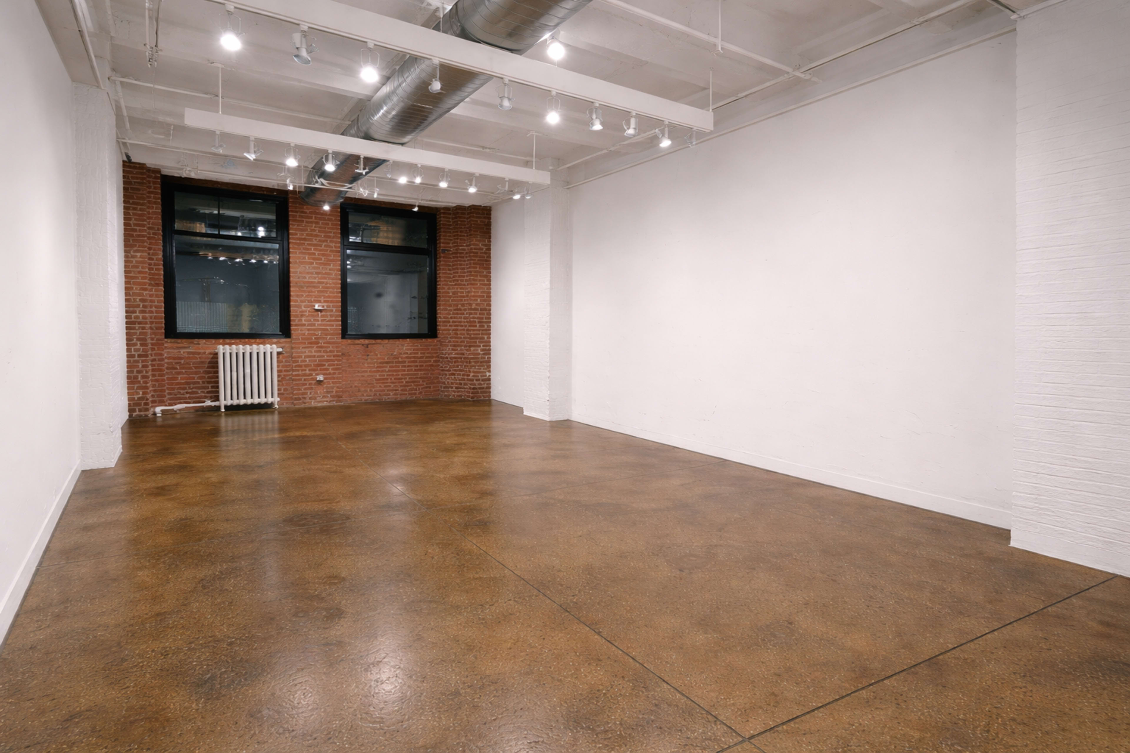 The image shows a spacious, empty room with polished concrete floors, exposed brick walls, and large windows.
