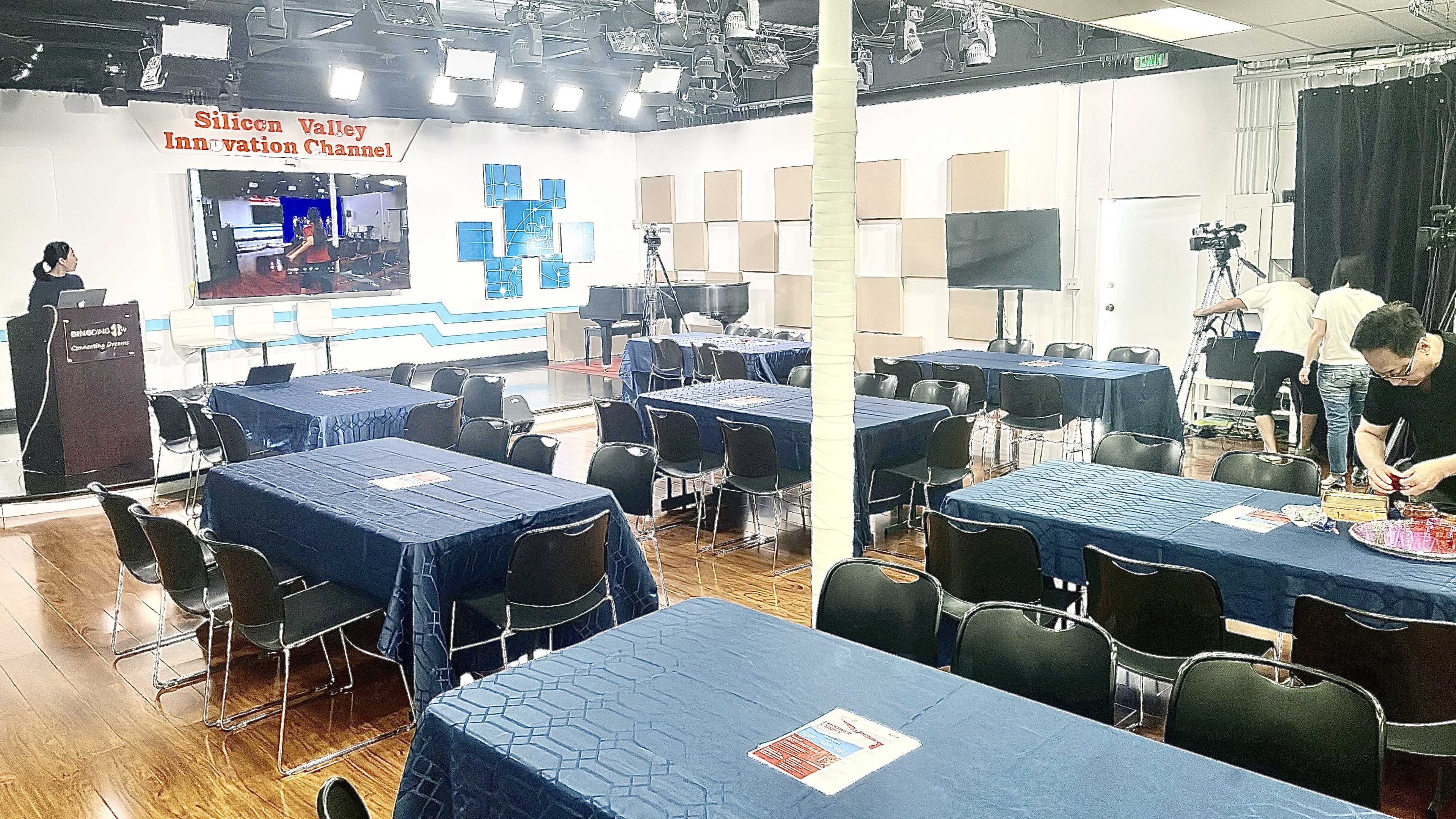 A spacious studio is set up for filming, featuring tables with blue tablecloths and various cameras positioned around the room.