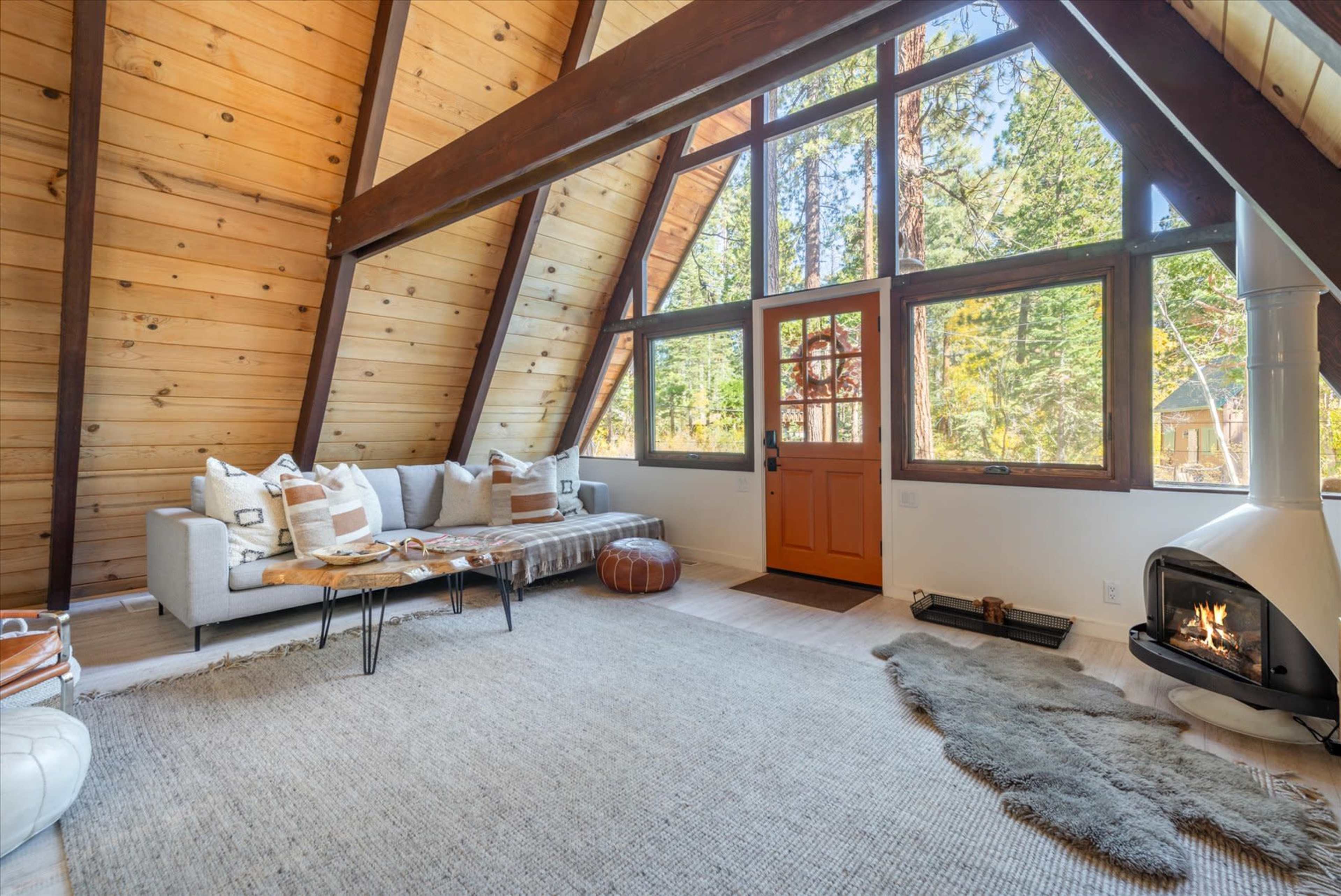 The image shows a rustic A-frame living room with large windows, a wooden beam ceiling, a cozy sofa, and a modern fireplace.