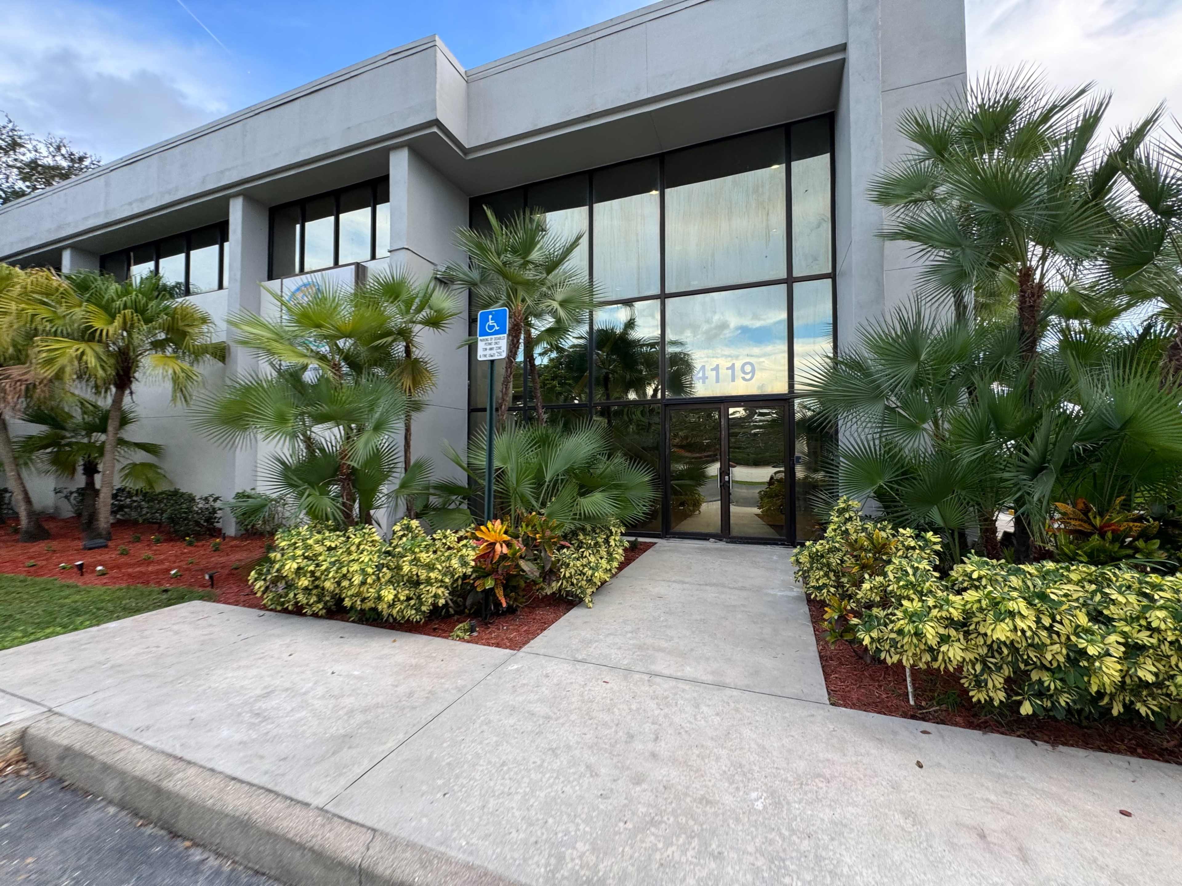 The image shows the entrance of a modern building with large glass windows, surrounded by landscaped plants and a pathway leading to the door.