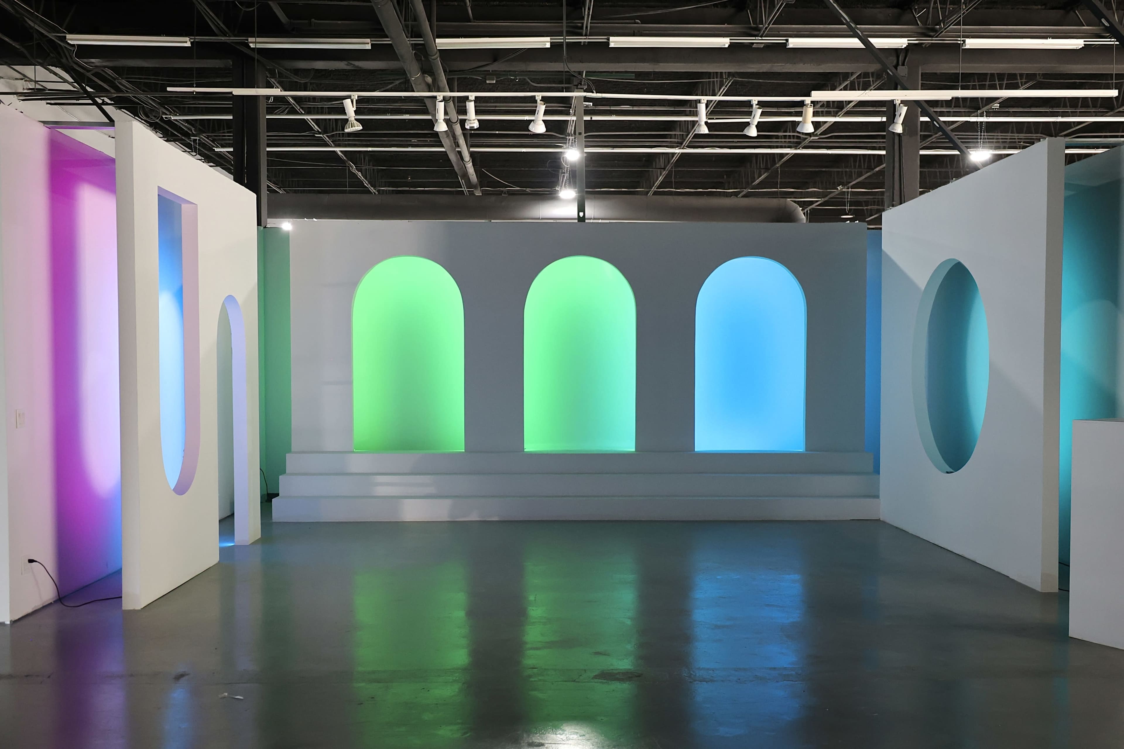 The image shows a modern art installation featuring several illuminated arches in various colors set against a white background.