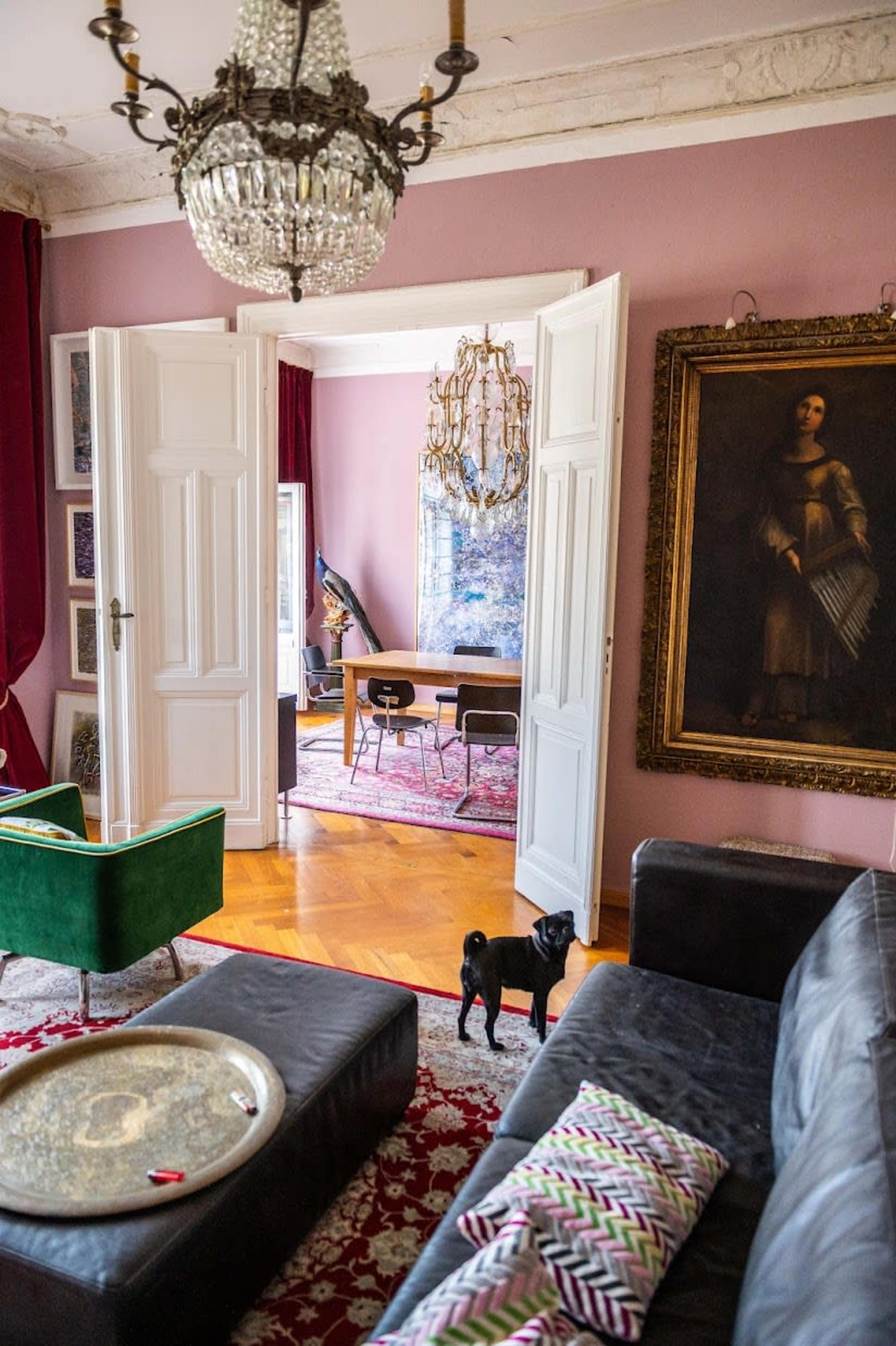A living room features a black dog standing near a dark sofa, with an ornate chandelier above and a doorway leading to another room with a chandelier and chairs.