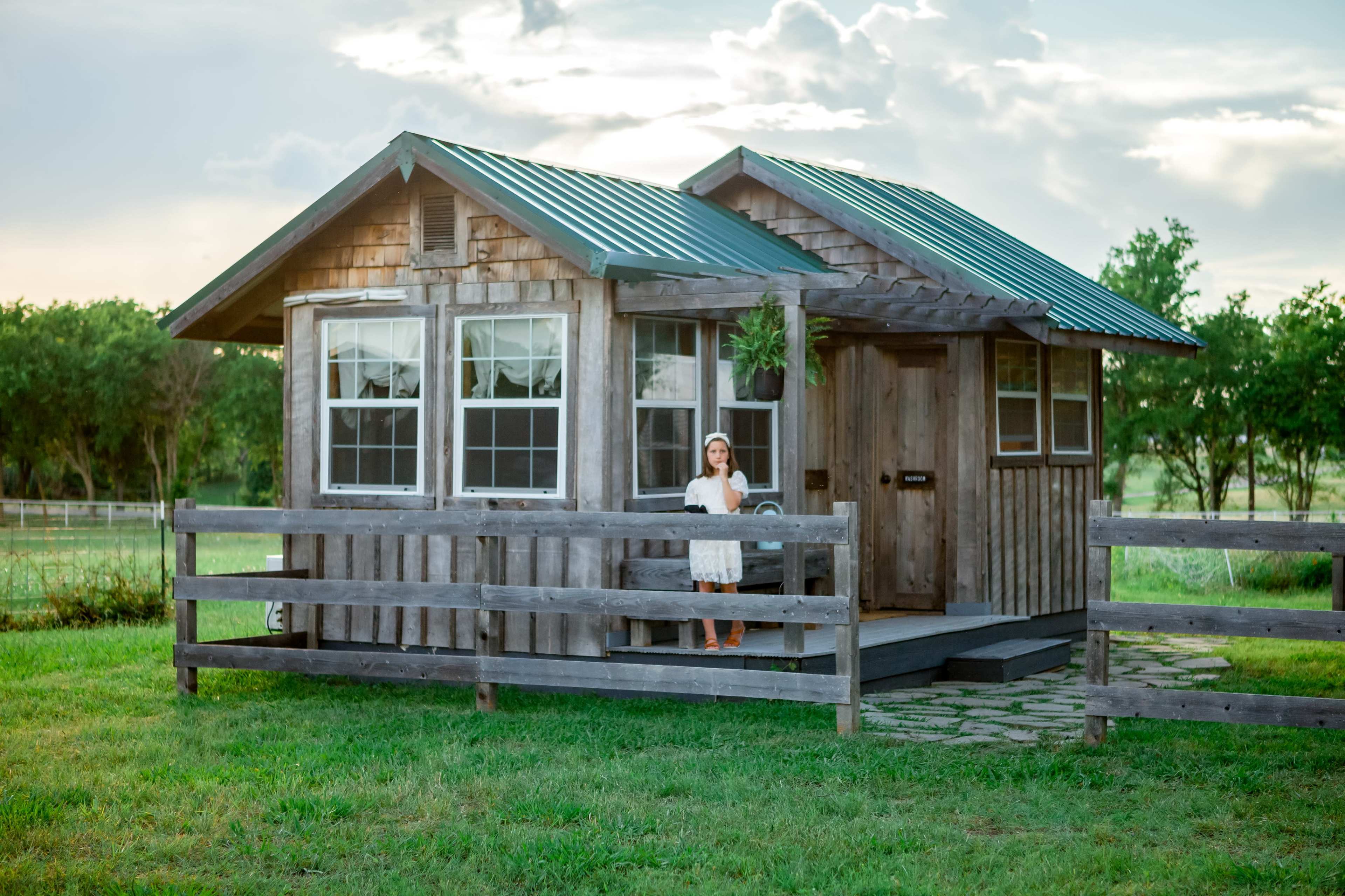 A woman stands on the porch of a small wooden cabin with a green metal roof, surrounded by a grassy area and trees in the background.