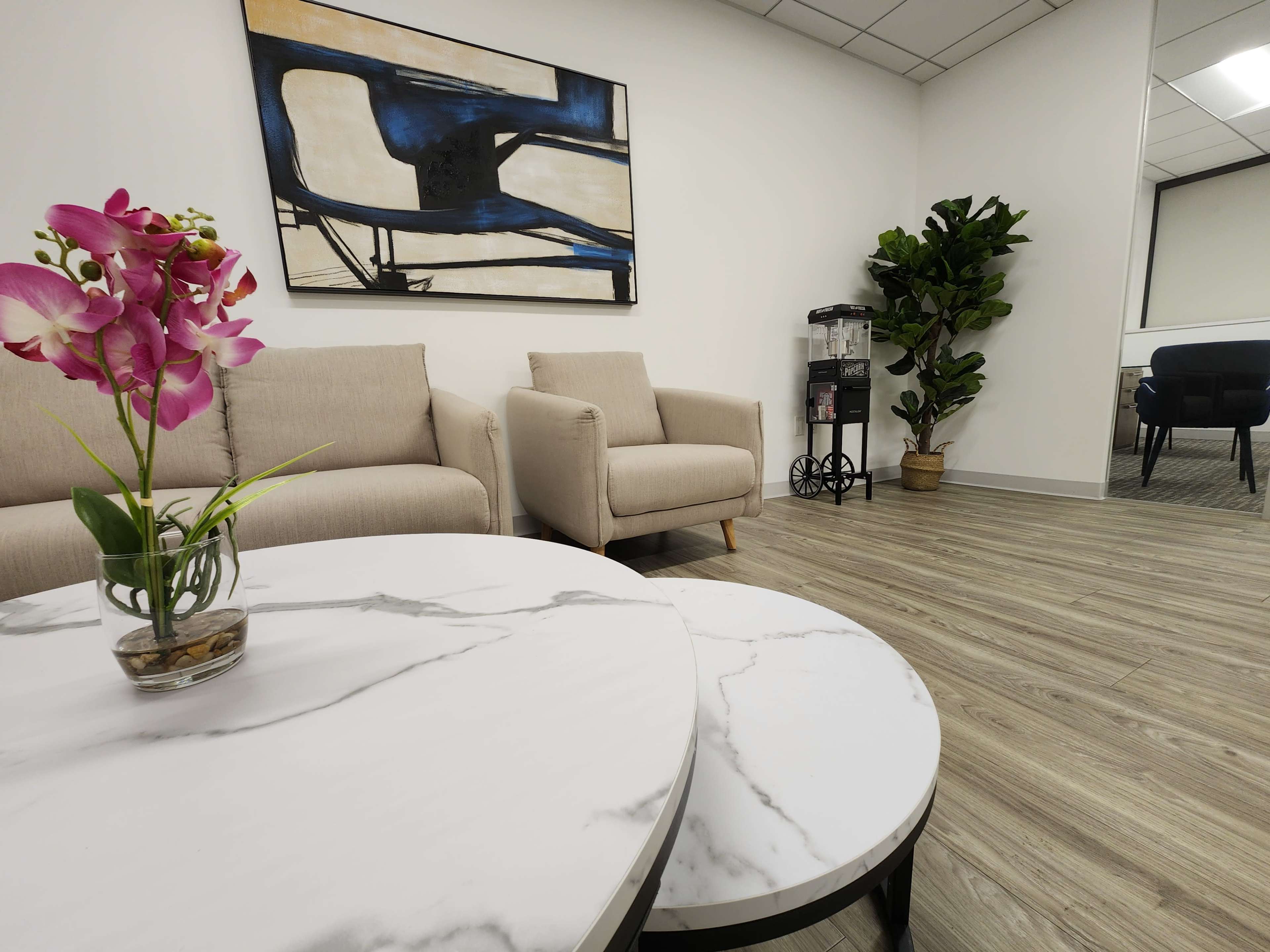 The image shows a modern waiting area with two beige sofas, a marble coffee table, a potted plant, and a wall art piece in a well-lit room.