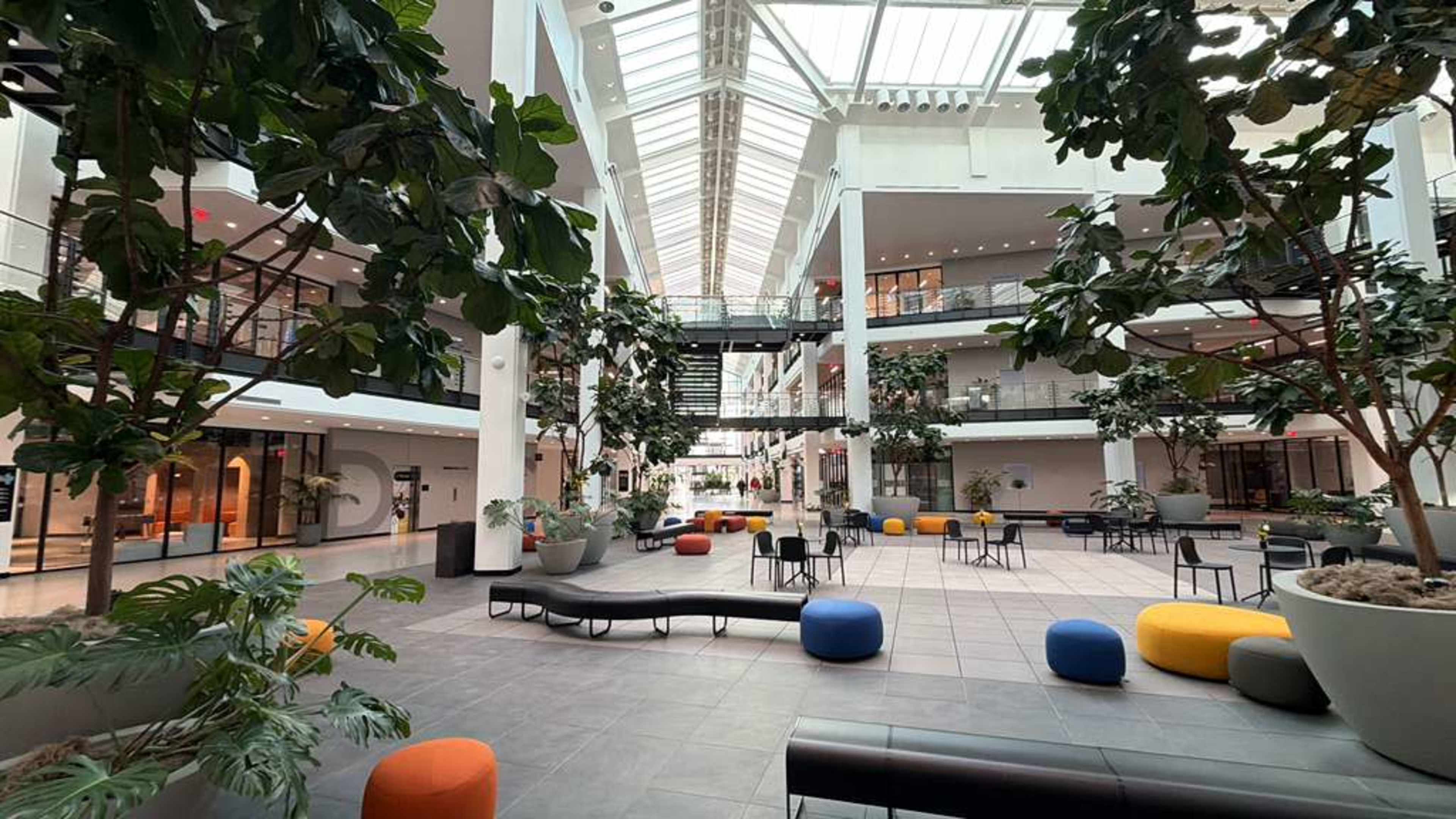 The image shows a modern indoor space with multiple levels, featuring large potted plants, various seating arrangements, and ample natural light from a glass roof.