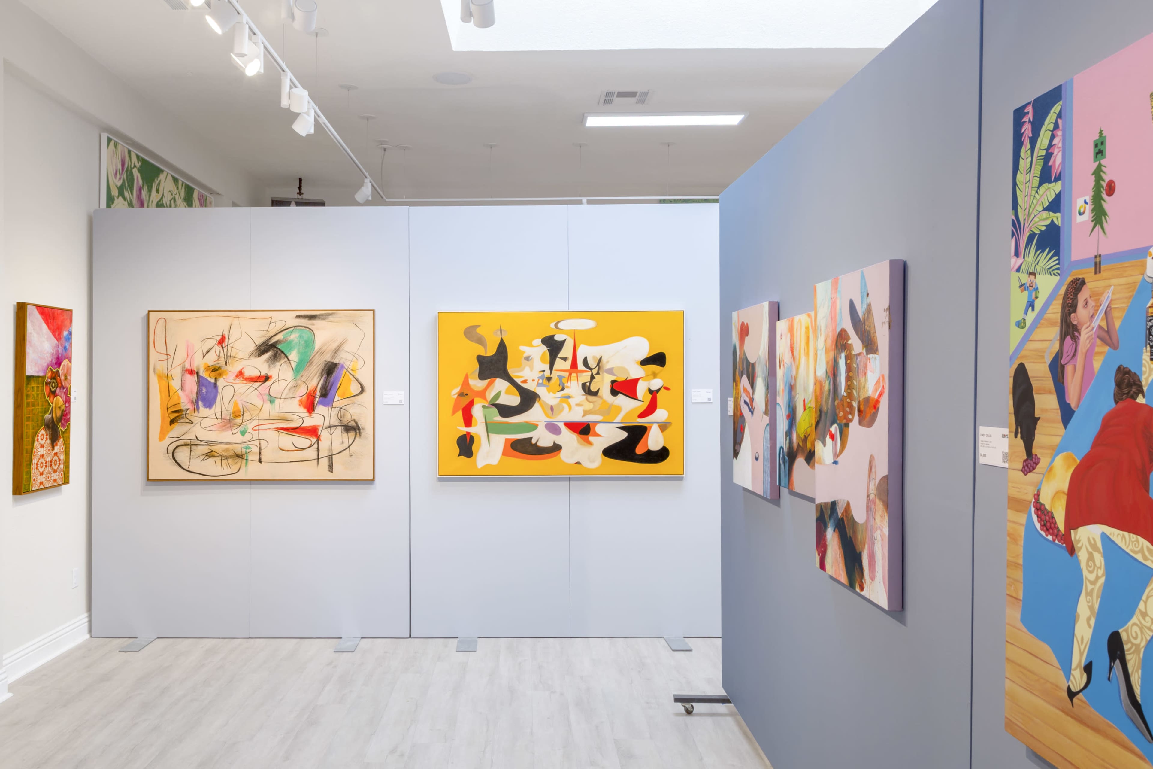 The image shows an art gallery featuring several colorful paintings displayed on light gray walls.