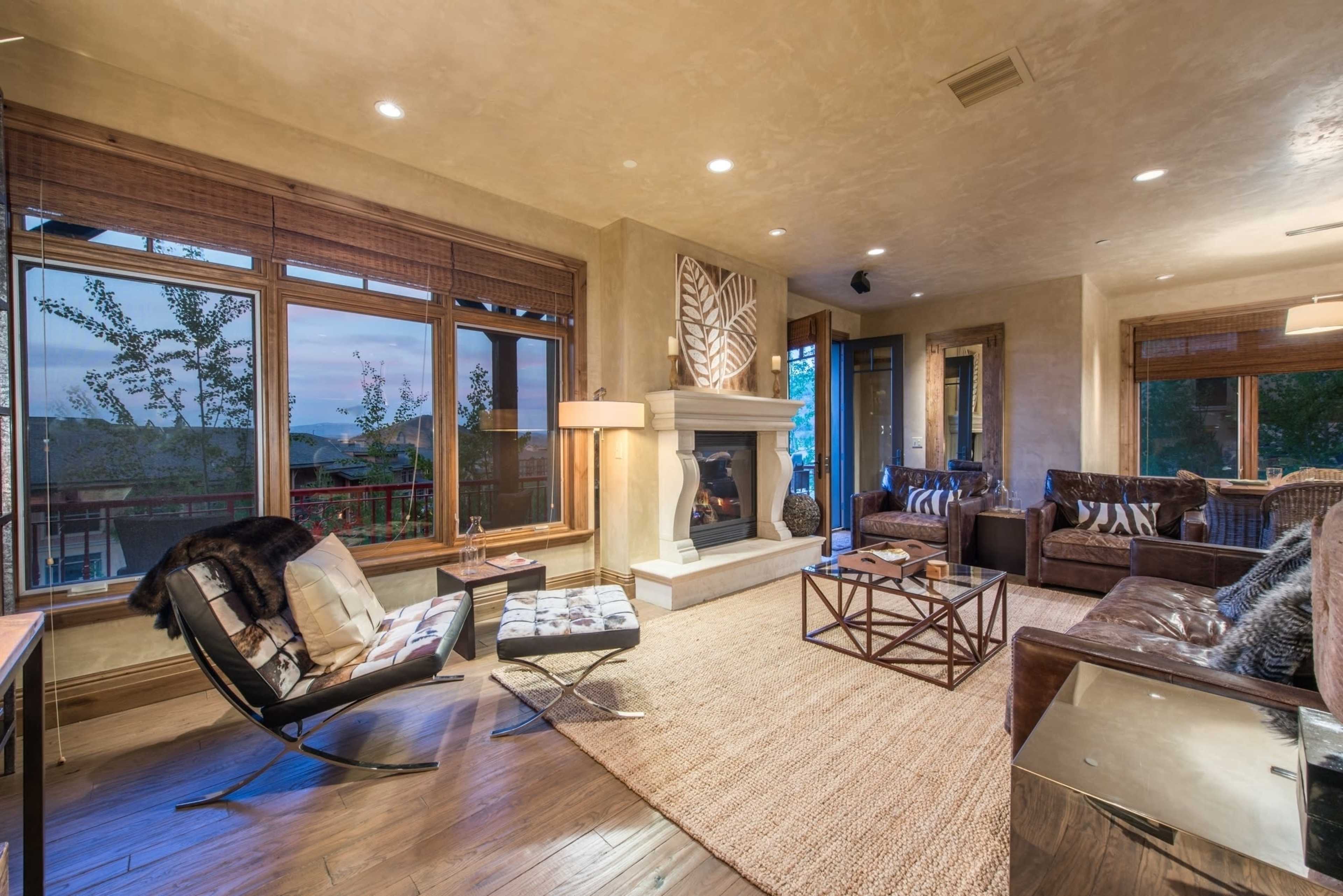 A cozy living room features a fireplace, large windows with views of trees, and a blend of modern and rustic furnishings.