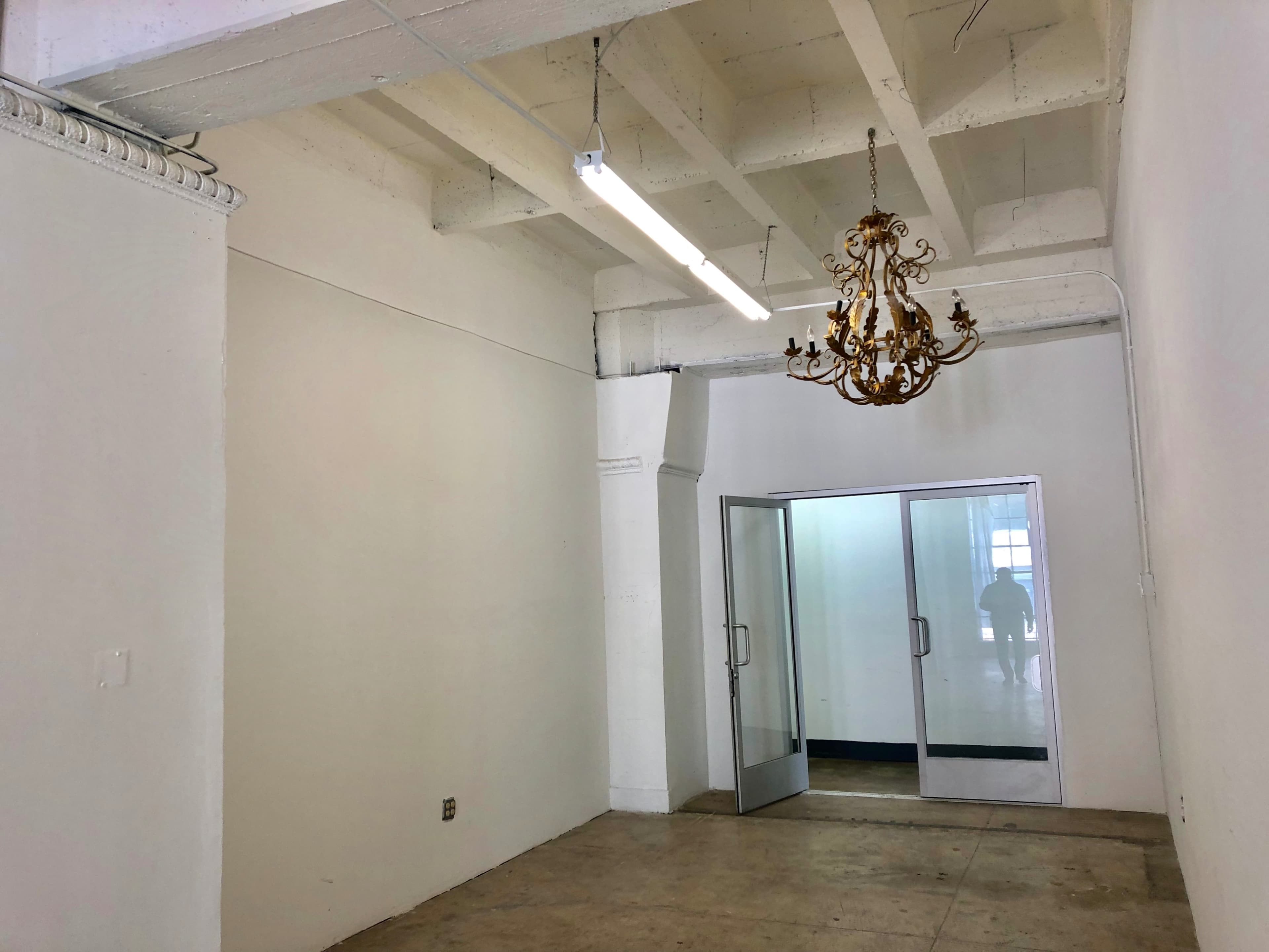 The image shows an empty room with a concrete floor, white walls, and a decorative chandelier hanging from the ceiling.
