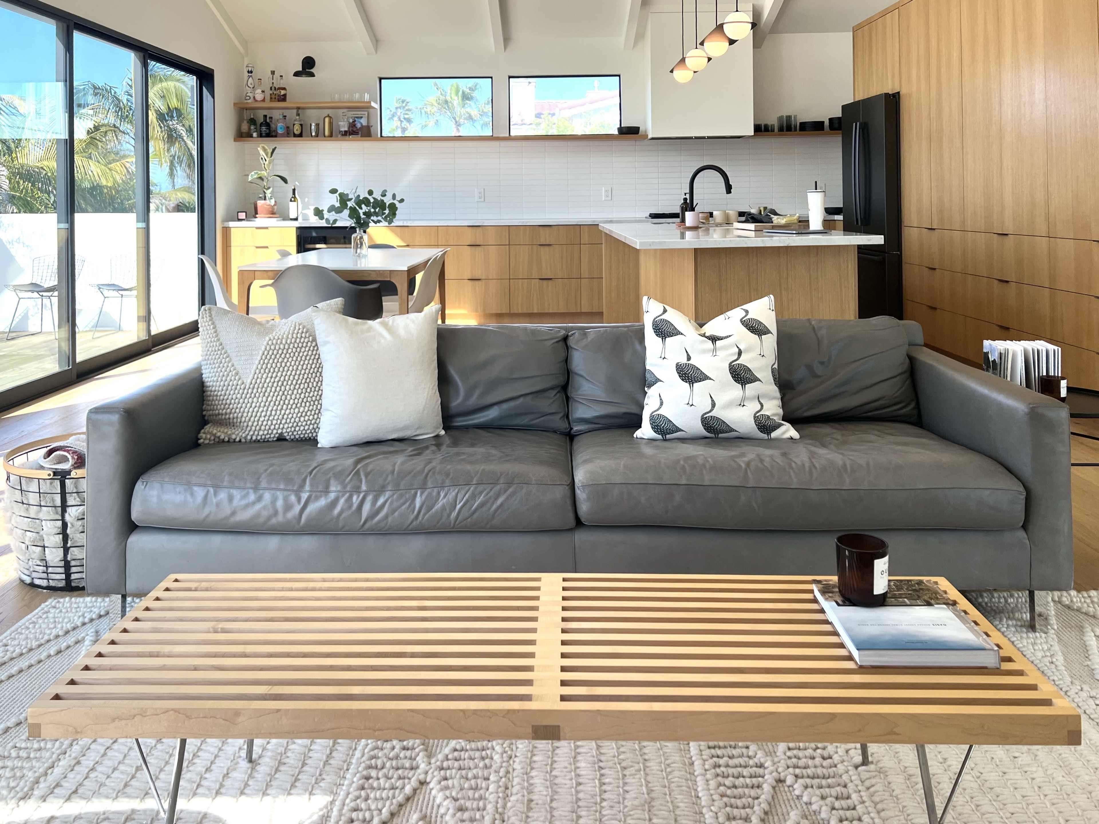 A modern living room features a gray leather sofa adorned with patterned pillows, a wooden coffee table, and an open kitchen in the background.