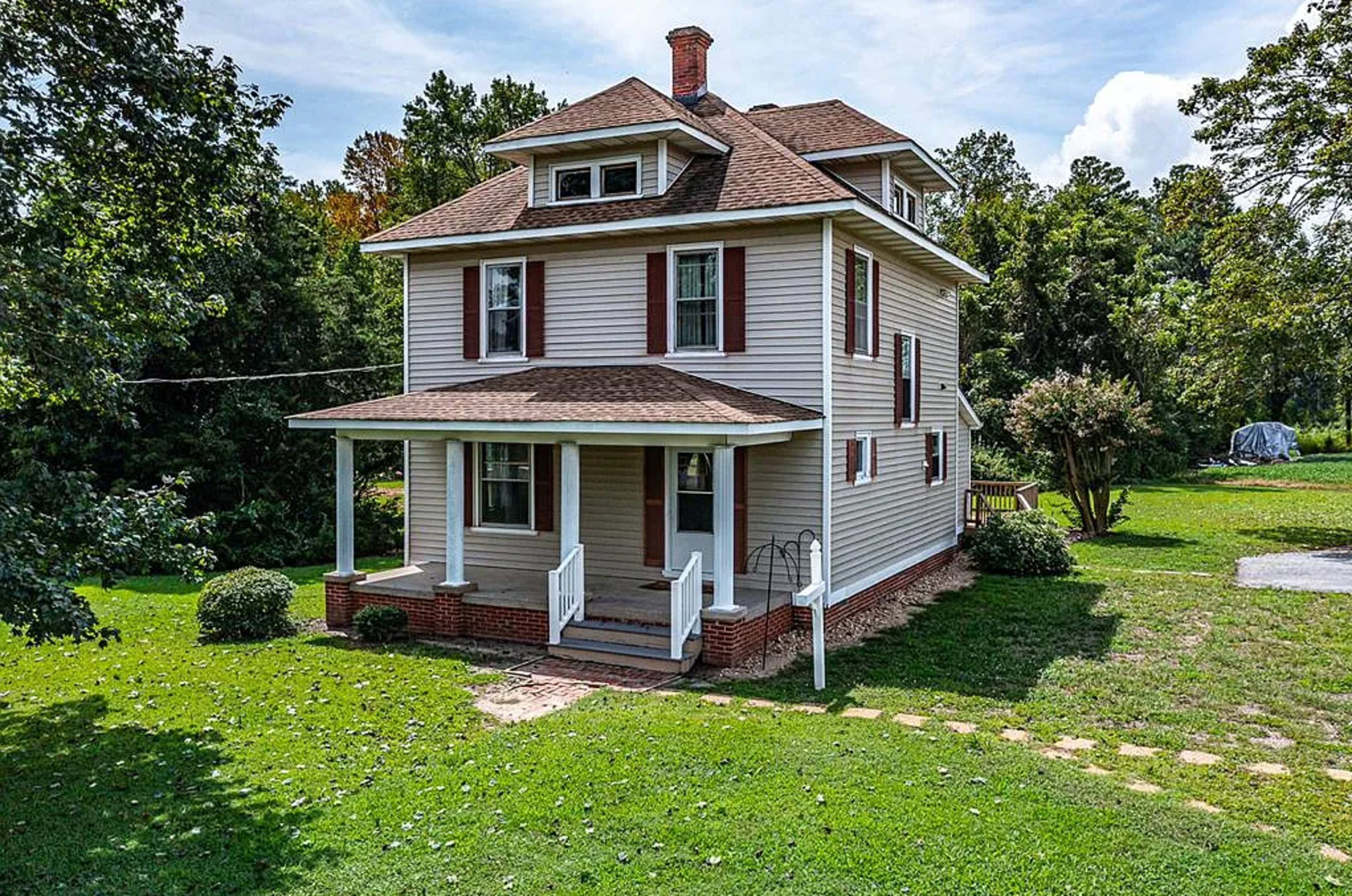 The image shows a three-story house with a front porch, surrounded by green grass and trees.