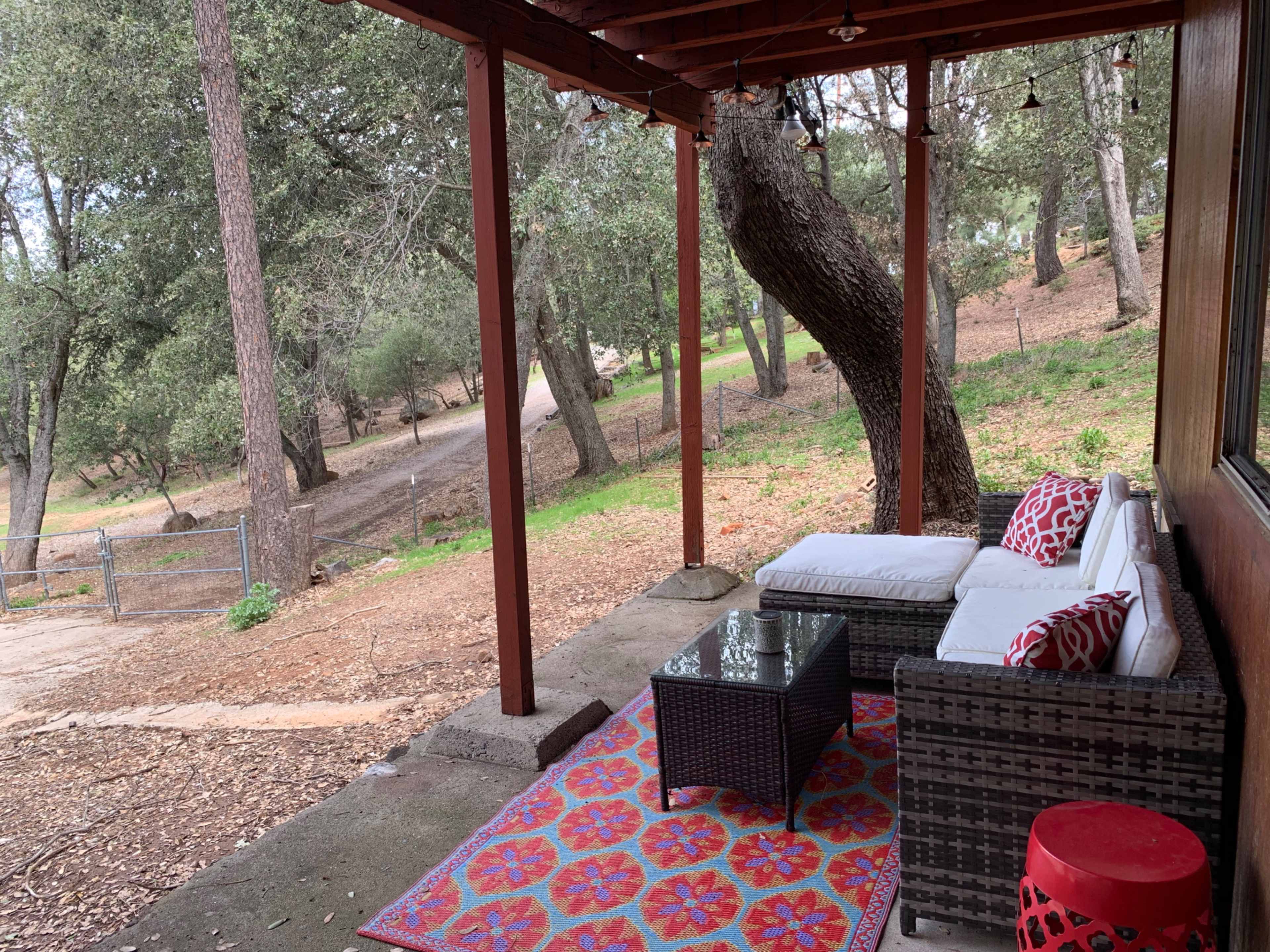 A covered porch features a seating area with a couch and glass coffee table, surrounded by trees and a gravel pathway.