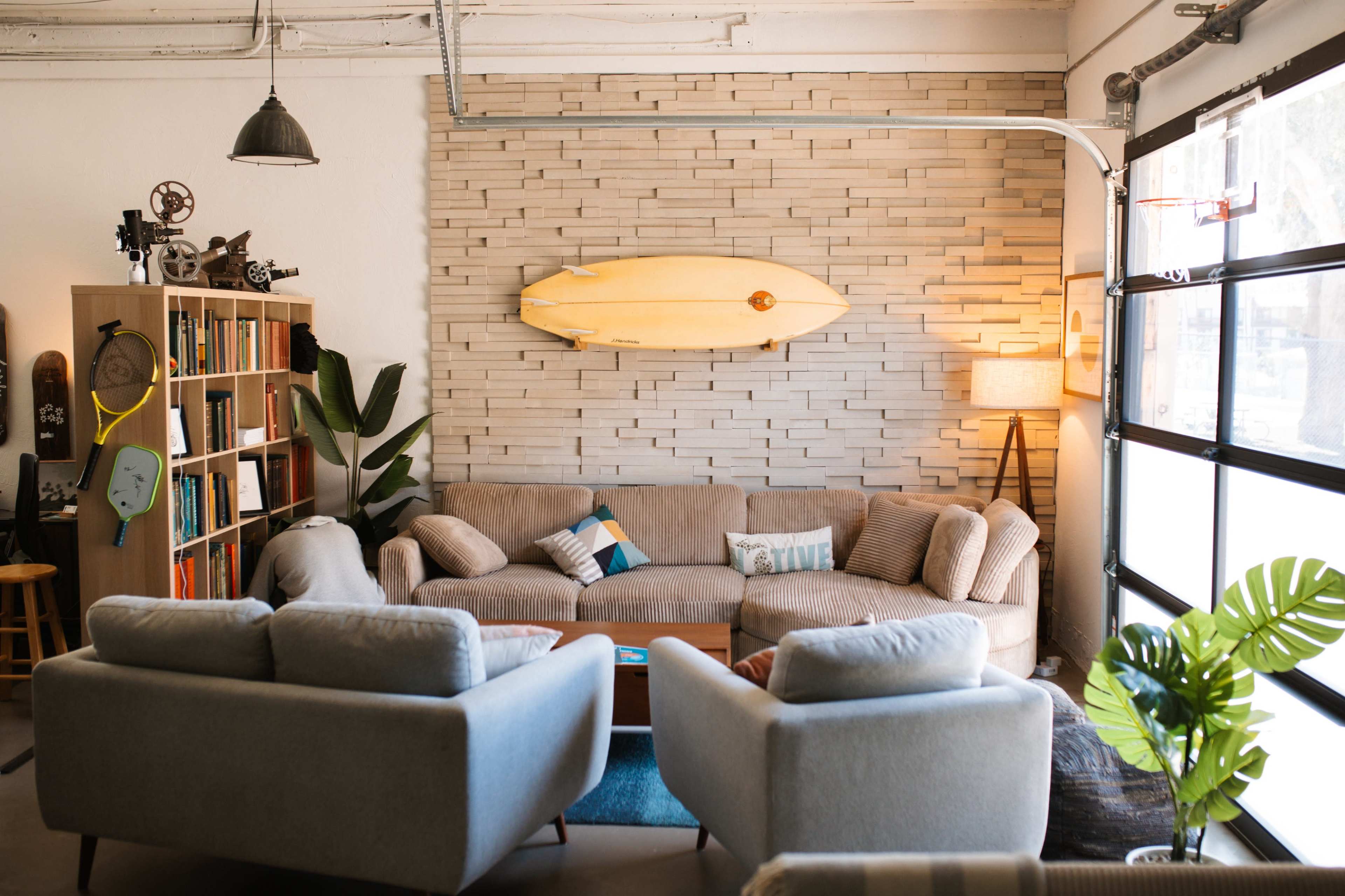 The image shows a modern living space featuring a beige couch, two light gray armchairs, a surfboard mounted on a textured wall, and a bookshelf filled with books.