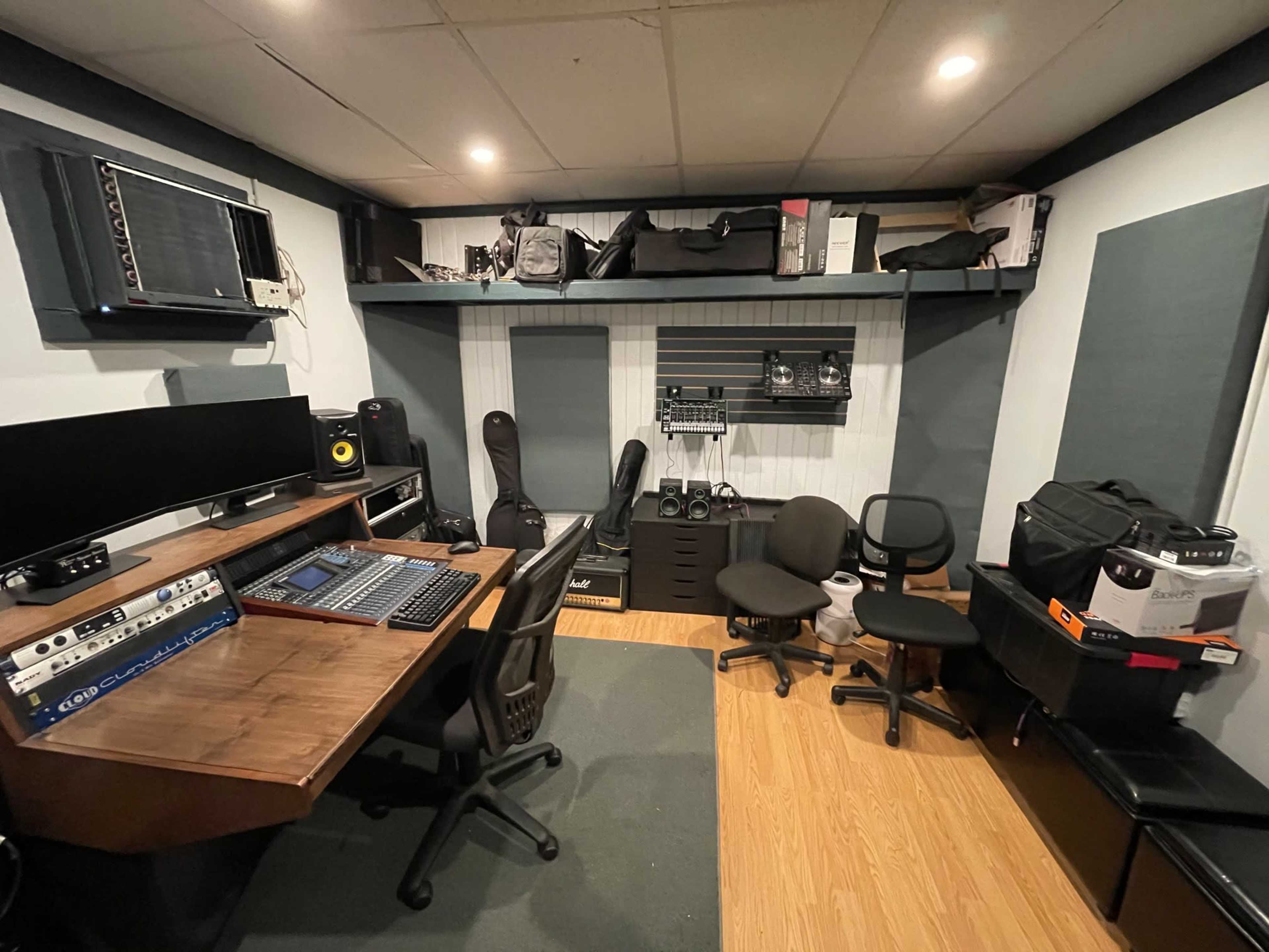 A compact audio recording studio with a wooden desk, computer monitors, various audio equipment, and storage shelves filled with bags and gear.