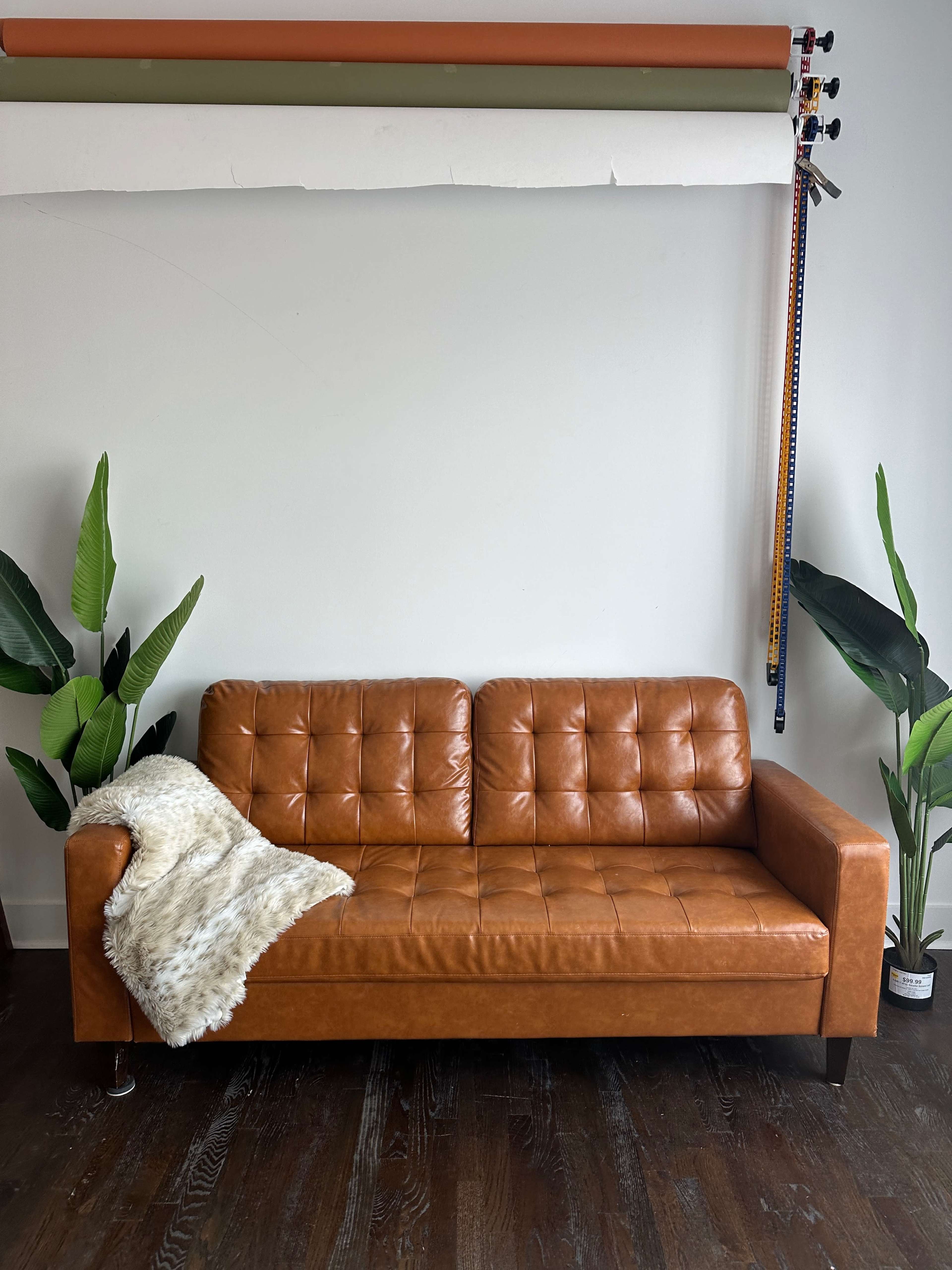 A brown leather sofa with tufted cushions is positioned against a wall, accompanied by decorative plants and a fur throw blanket.