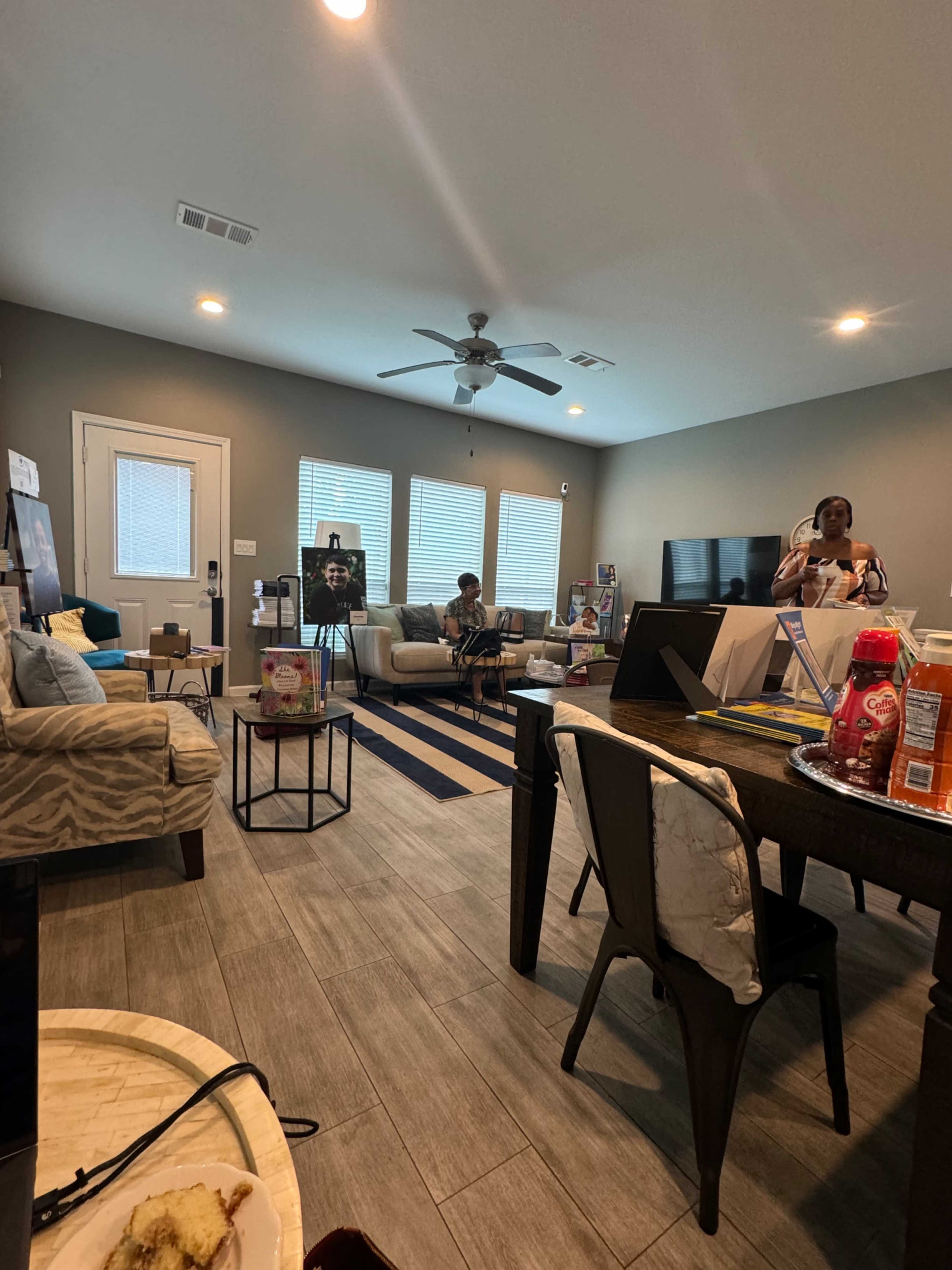 A living room features a dining table, seating areas, and multiple windows providing natural light.