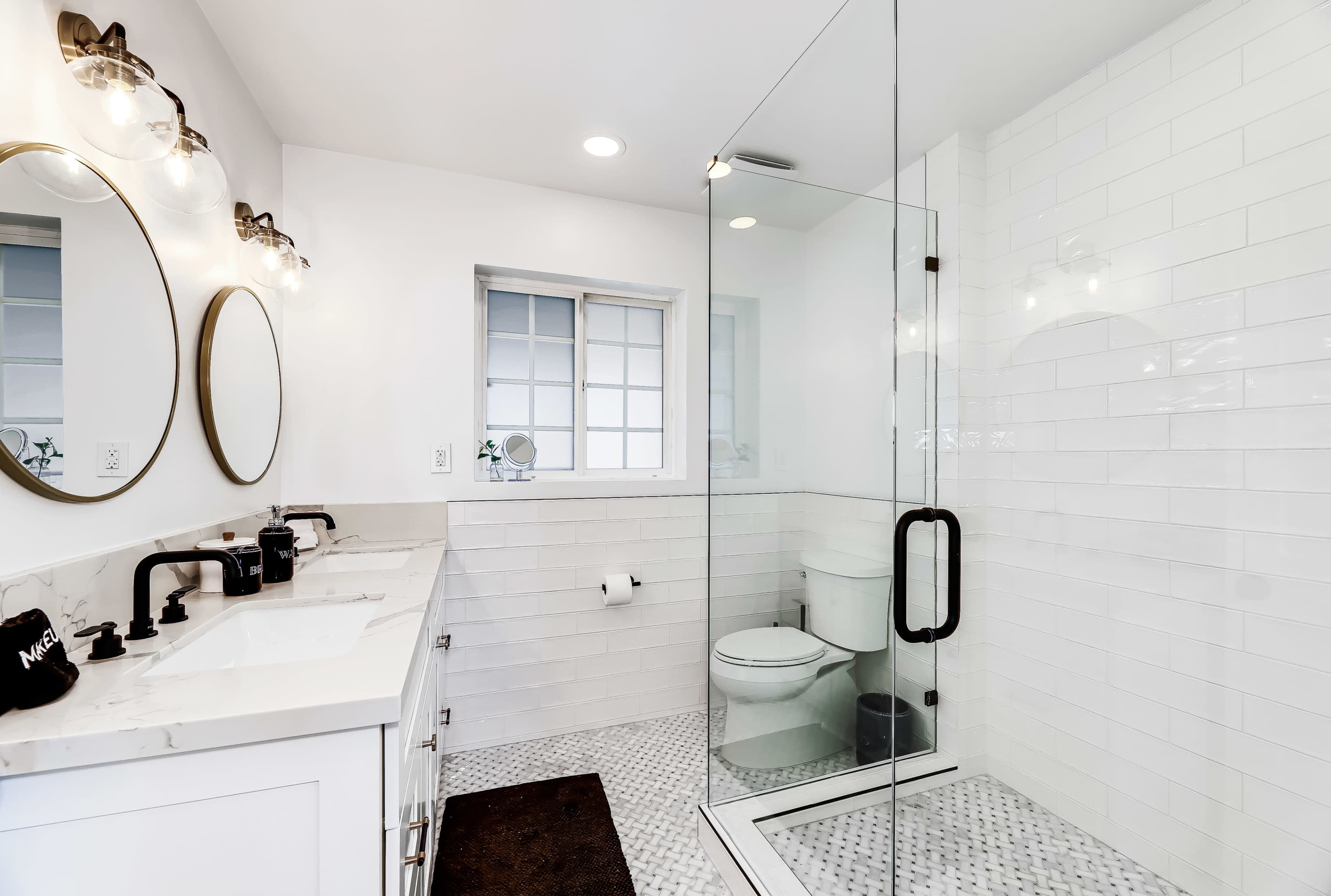 A modern bathroom features a walk-in shower, dual sinks with round mirrors, and white tiled walls.
