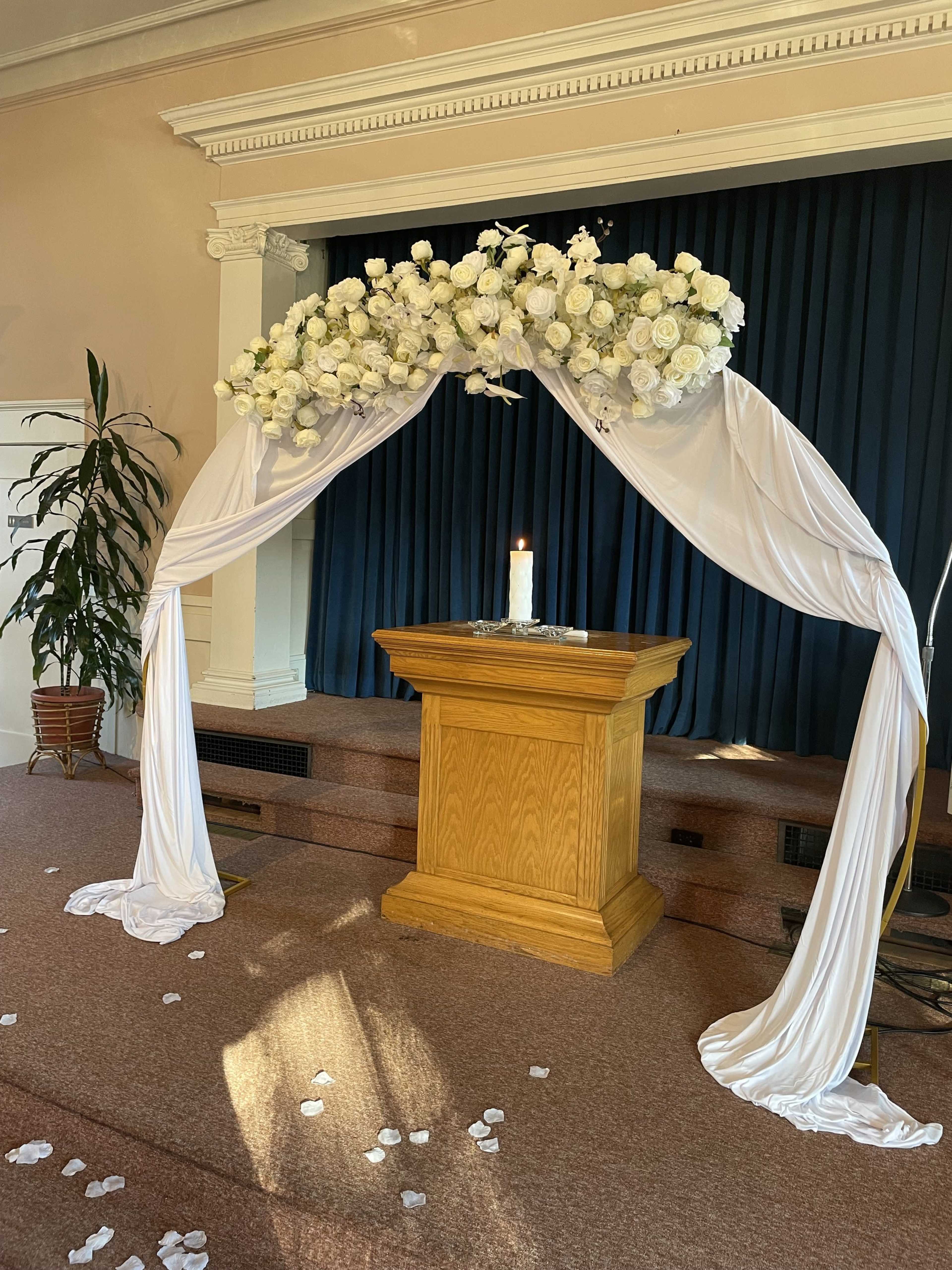 The image shows a decorated wedding altar with a floral arch, draped fabric, and a lit candle positioned on a wooden podium, with scattered petals on the floor.